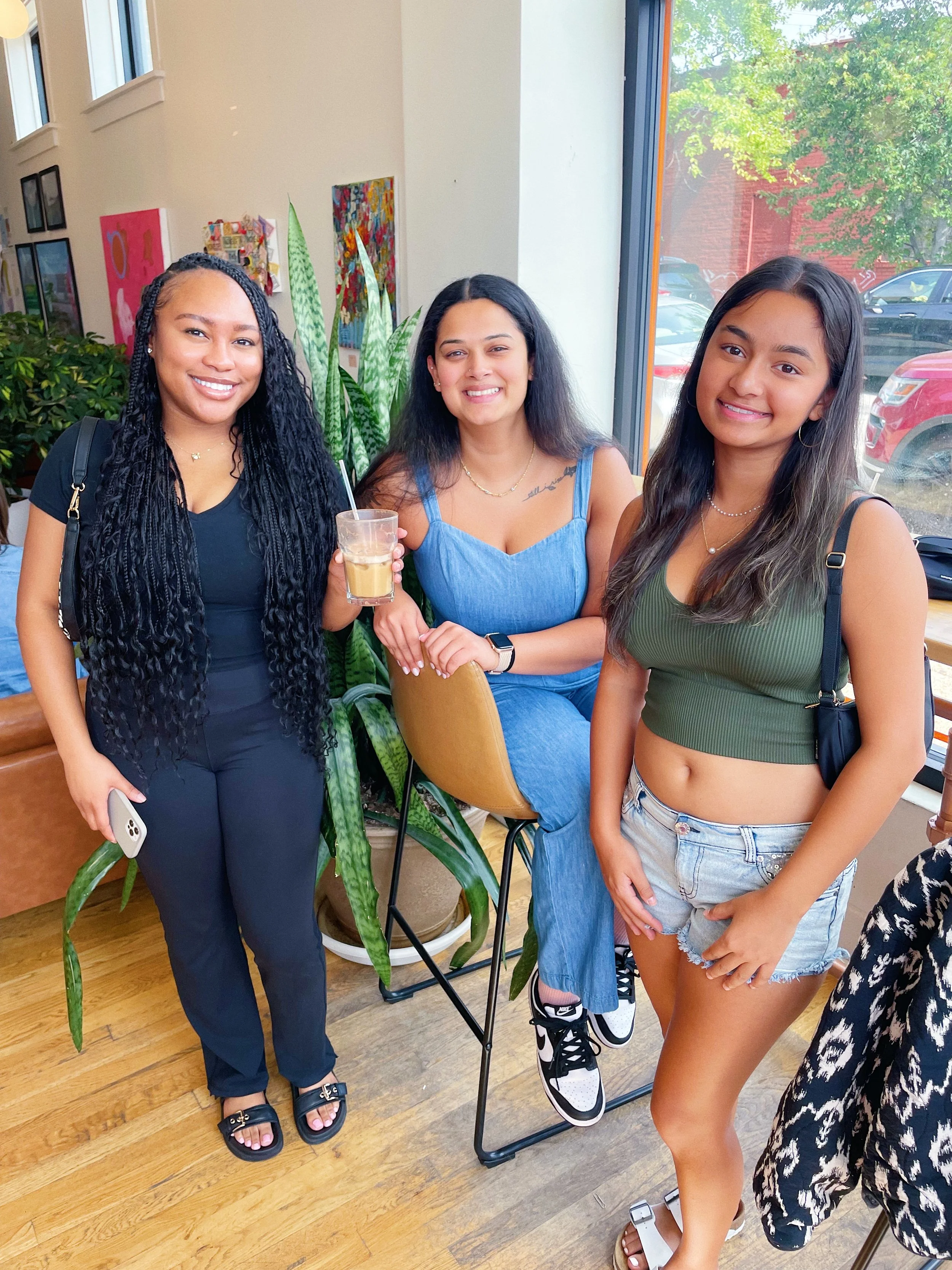 Three women smiling indoors near a large window, with plants and colorful artwork behind them. The woman on the left is holding a drink, the middle woman is sitting on a stool, and the woman on the right is standing. All are dressed casually.