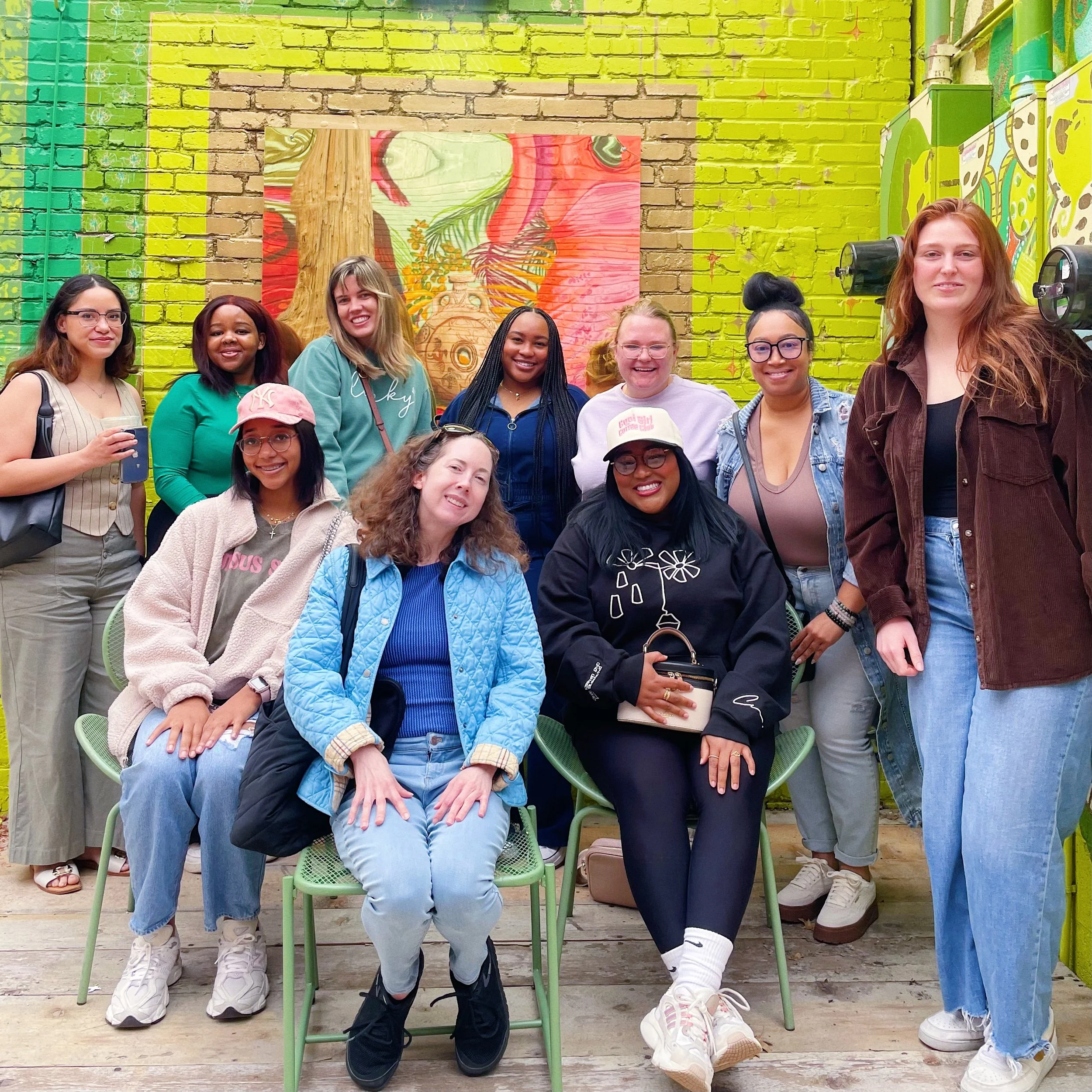 A diverse group of eleven women smiling and posing together indoors against a colorful, artistic background with a brick wall painted in bright yellow and green. Some women are sitting on green chairs, and others are standing behind them, dressed cas
