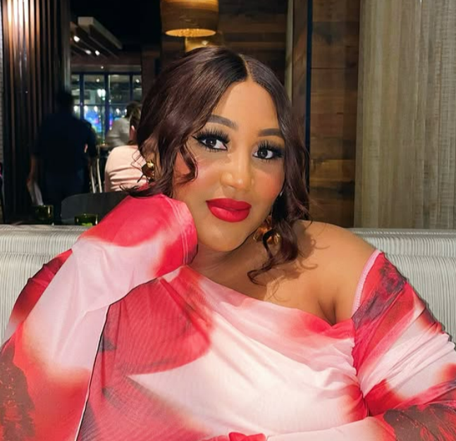 A woman with dark wavy hair and makeup, wearing a red and pink off-shoulder dress, sitting at a restaurant or cafe with a warm, modern interior.