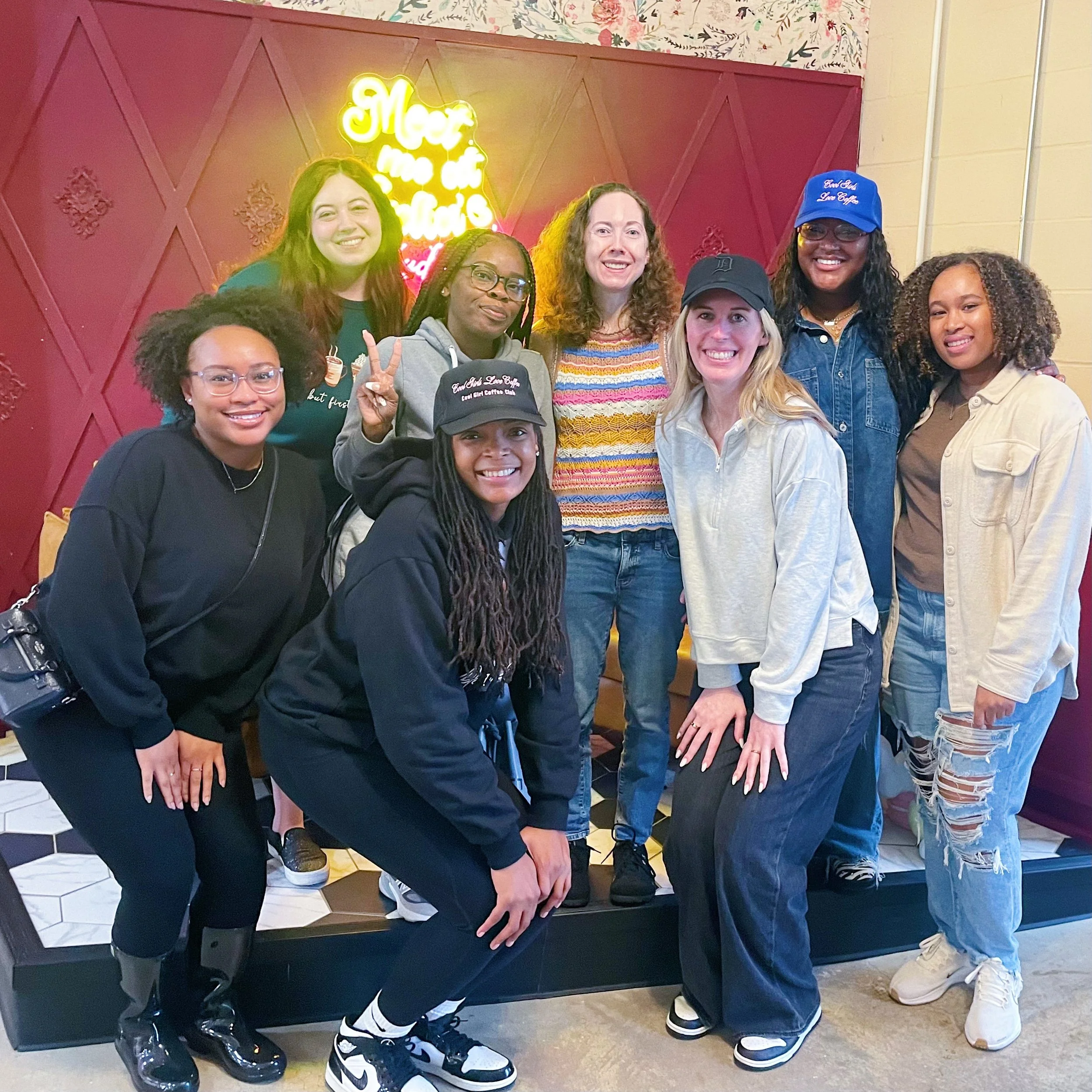 Group of nine women smiling and posing together in a restaurant or cafe with a red wall background and neon sign that reads 'More than a meal'.