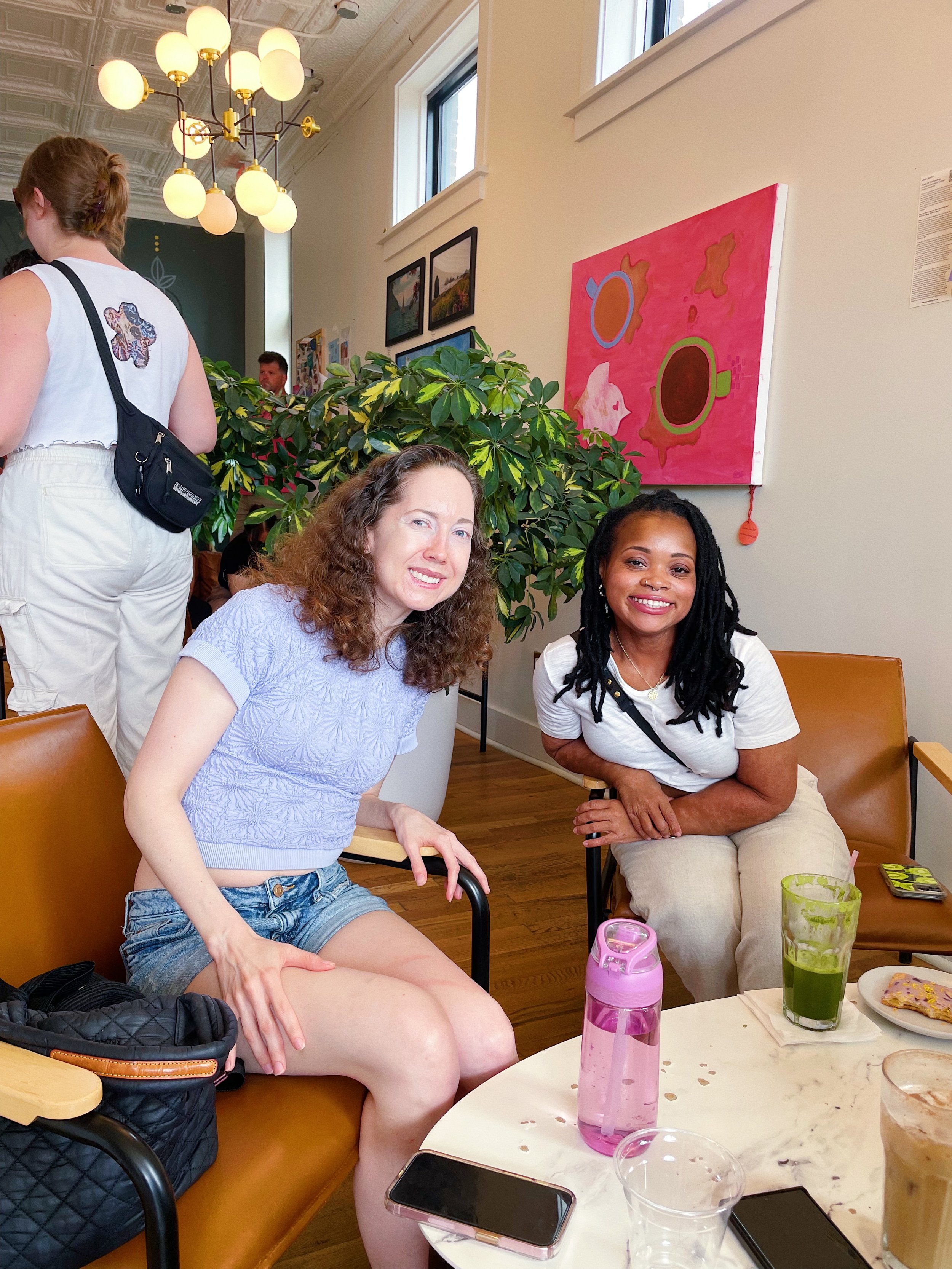 Two women sitting at a table in a cafe, smiling at the camera. One has curly brown hair and is wearing a light purple top and denim shorts. The other has black dreadlocks and is wearing a white t-shirt and beige pants. On the table are drinks, a phon