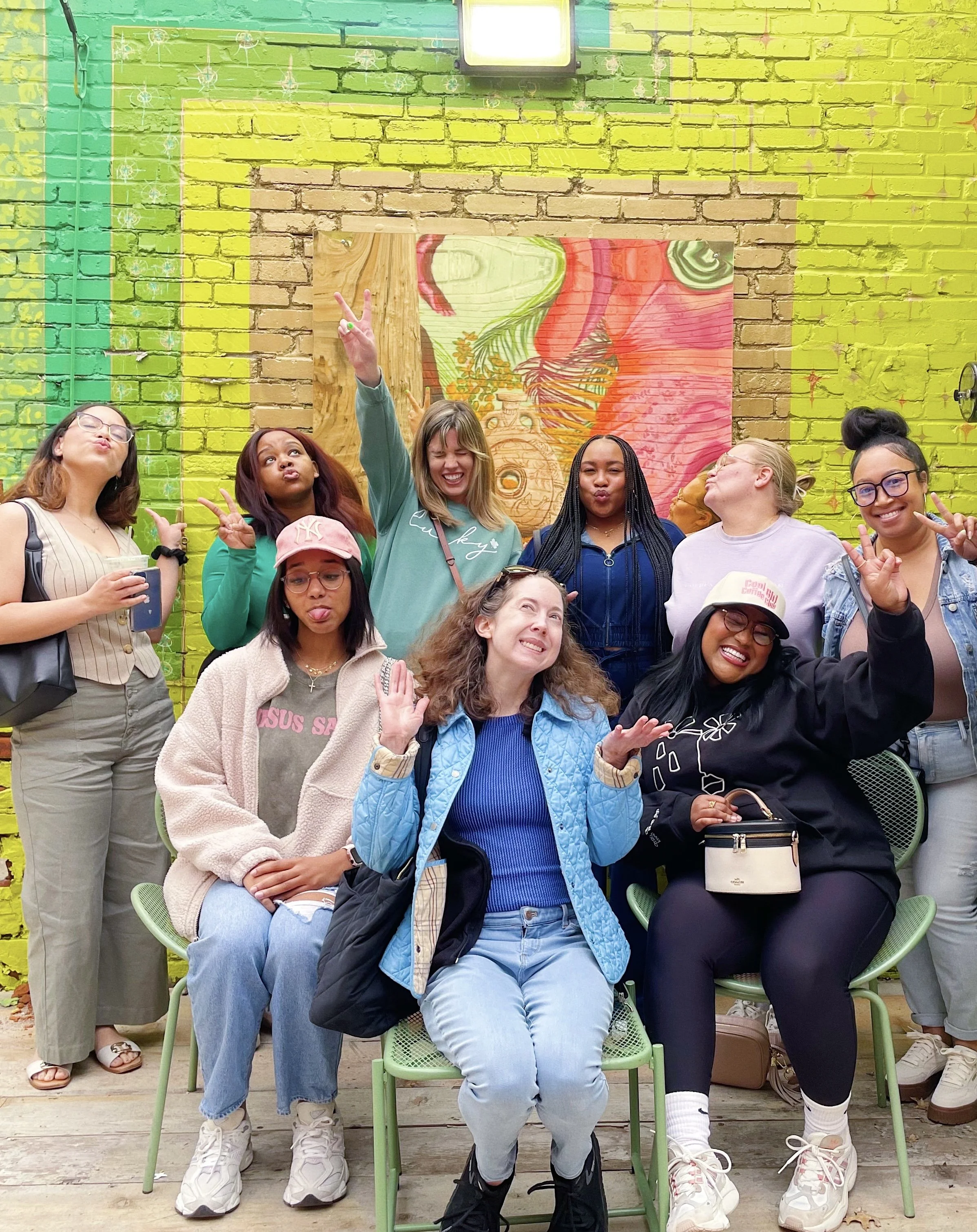 A group of nine women posing for a photo in front of a colorful brick wall with green, yellow, and orange colors and a large, abstract artwork in the background. Some women are making peace signs, and one is sticking out her tongue.