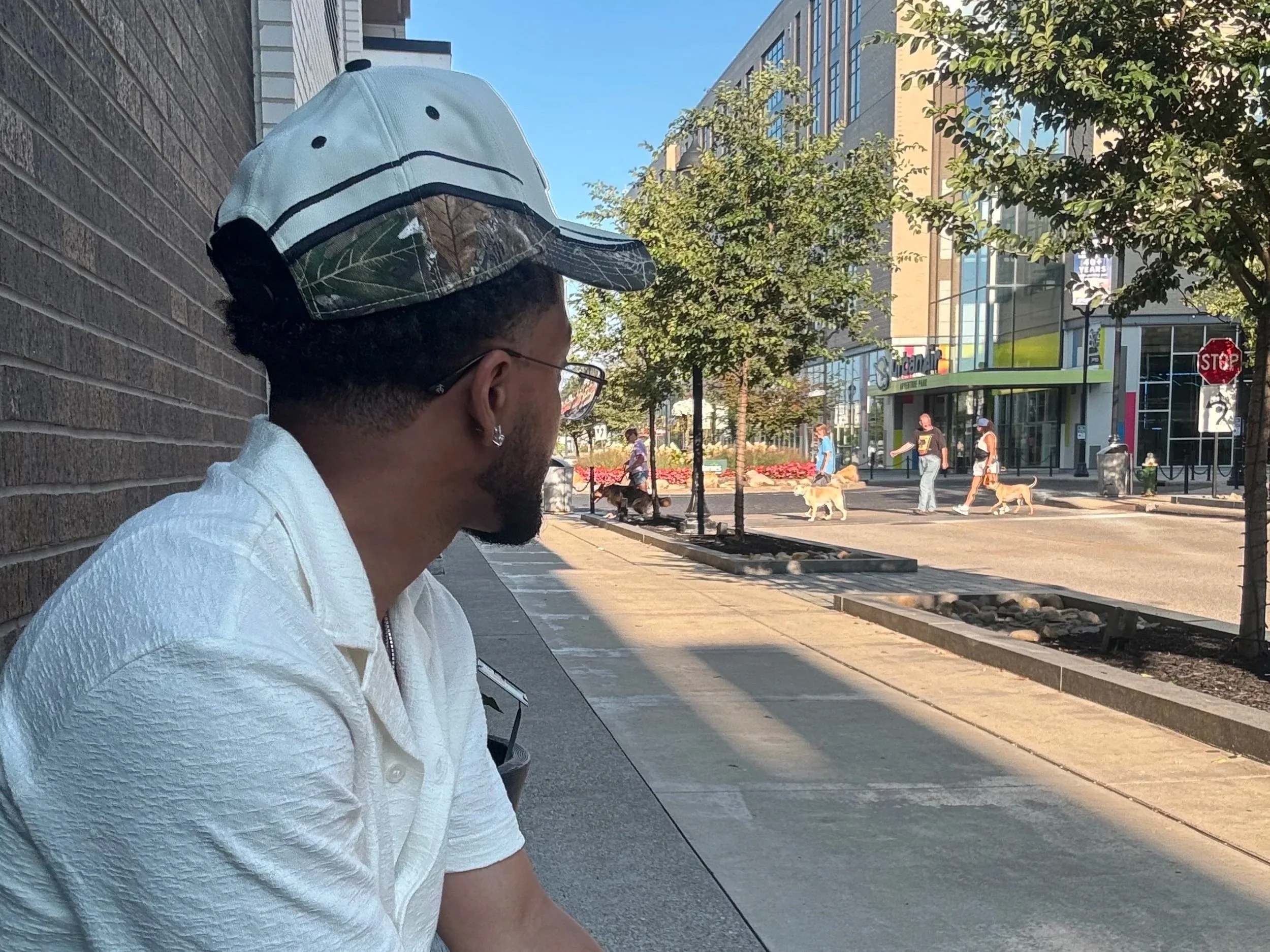 A man sitting on a bench on a city sidewalk, wearing a white shirt, glasses, earrings, and a cap decorated with leaves. In the background, people walking dogs across the street, modern buildings, and a stop sign are visible.