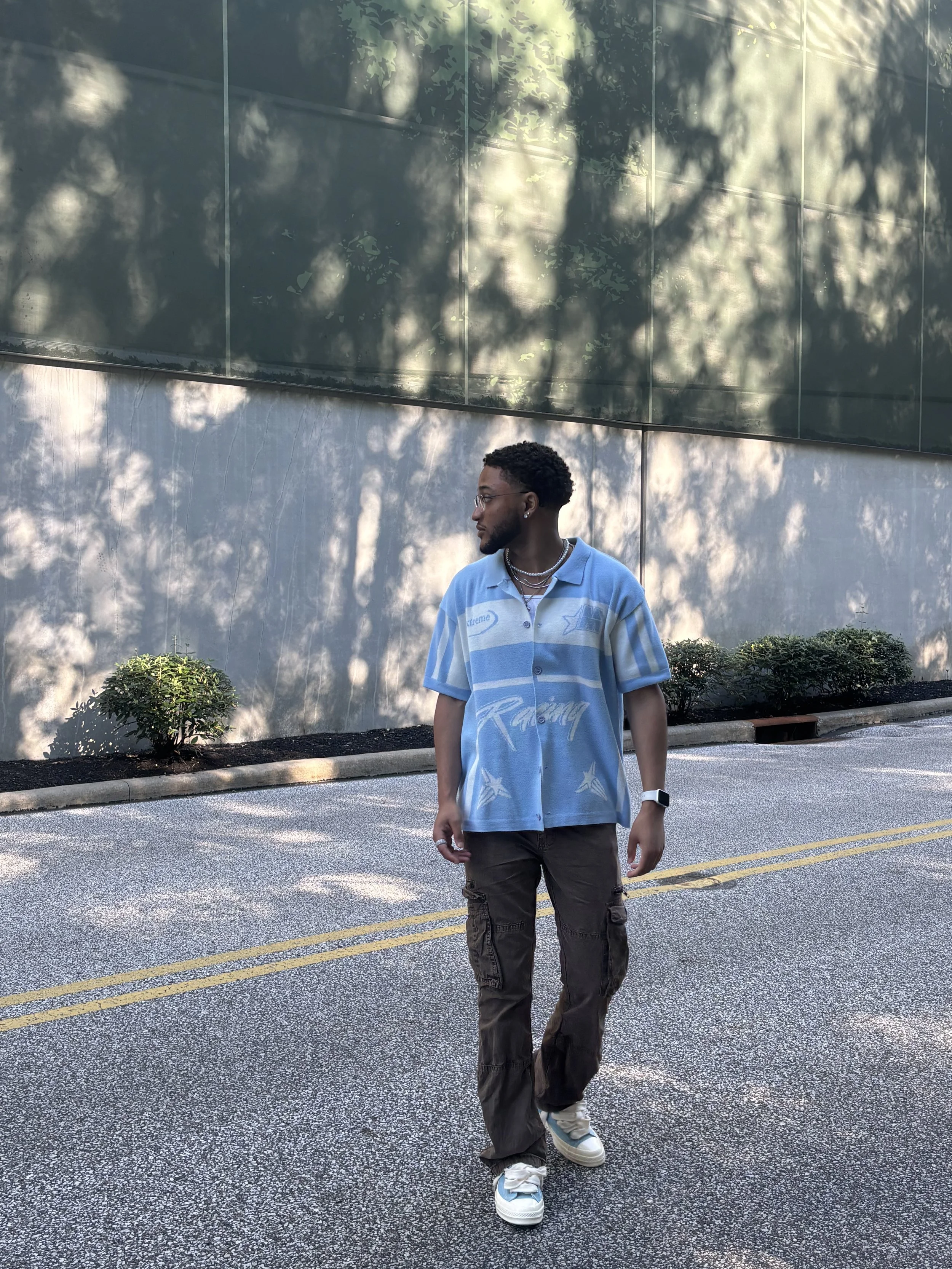 A young man with dark curly hair and glasses standing on a street with a large reflective glass building behind him. Shadows of trees are cast on the building. The man is wearing a light blue patterned oversized racing shirt, brown cargo pants, white sneakers, a wristwatch, and layered necklaces.