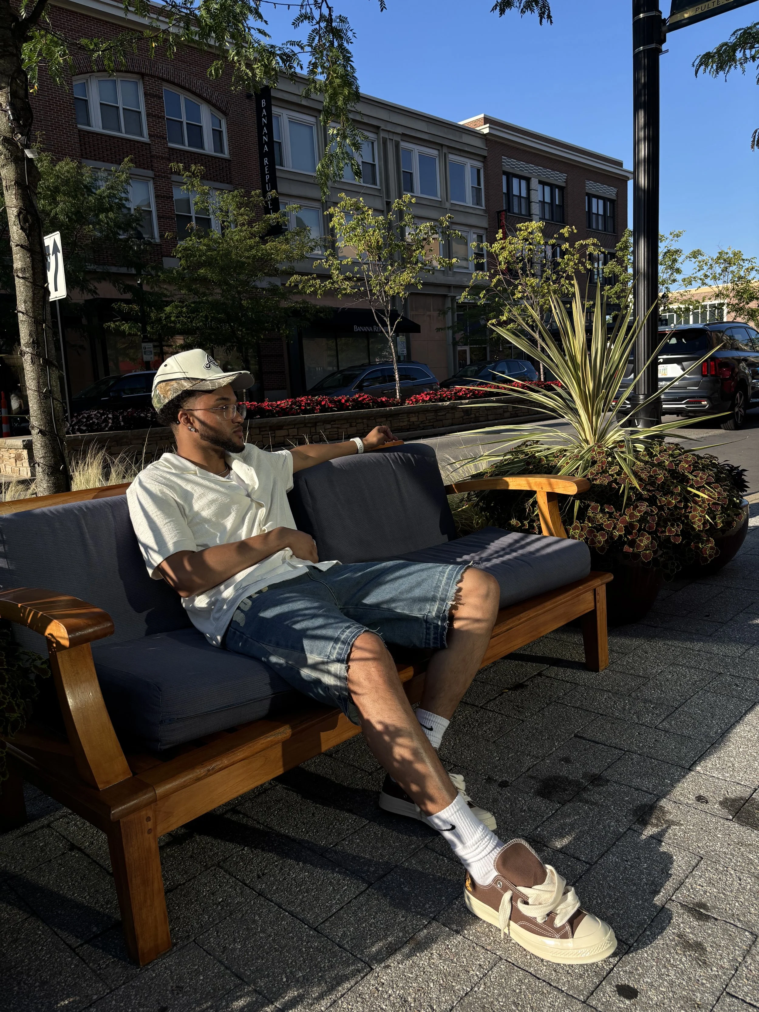 A young man sitting on a wooden bench outdoors, wearing a light-colored shirt, denim shorts, white socks, and sneakers, with a cap and sunglasses, with his left arm resting on the back of the bench. He is in shadow while the background features a storefront, trees, parked cars, and a clear blue sky.