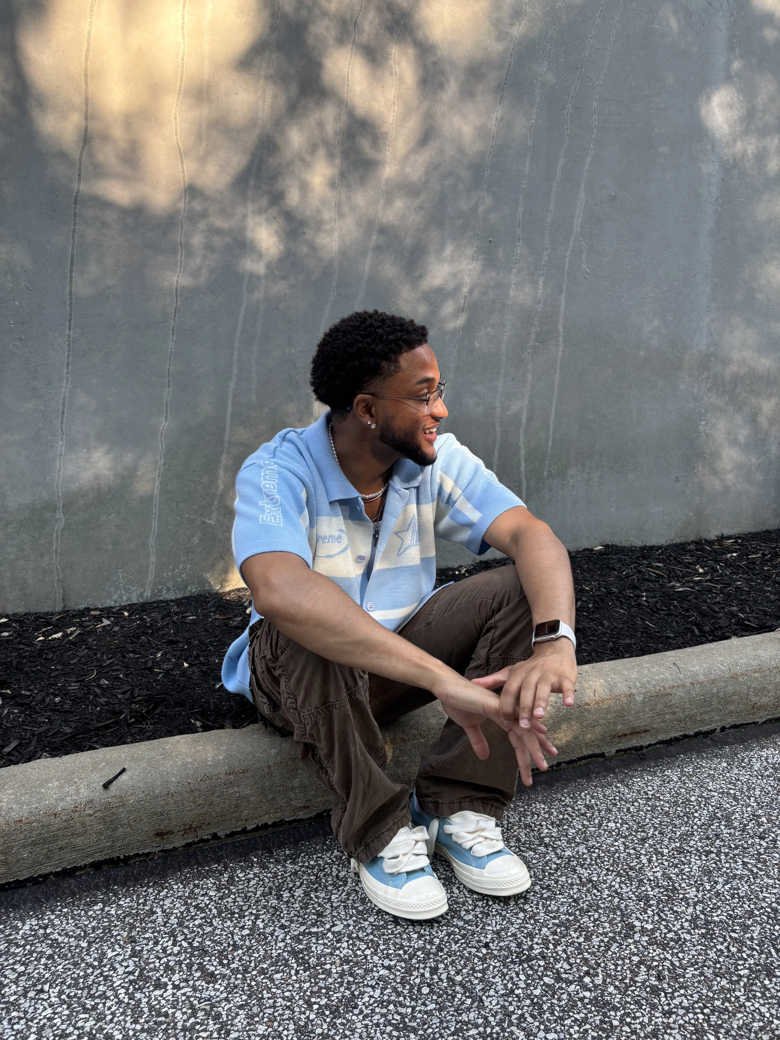 A young man with glasses, curly hair, and jewelry, sitting on a curb against a concrete wall with a cloud reflection, smiling and looking to his left, wearing a blue striped polo shirt, brown pants, white and blue sneakers, a watch, and a chain.