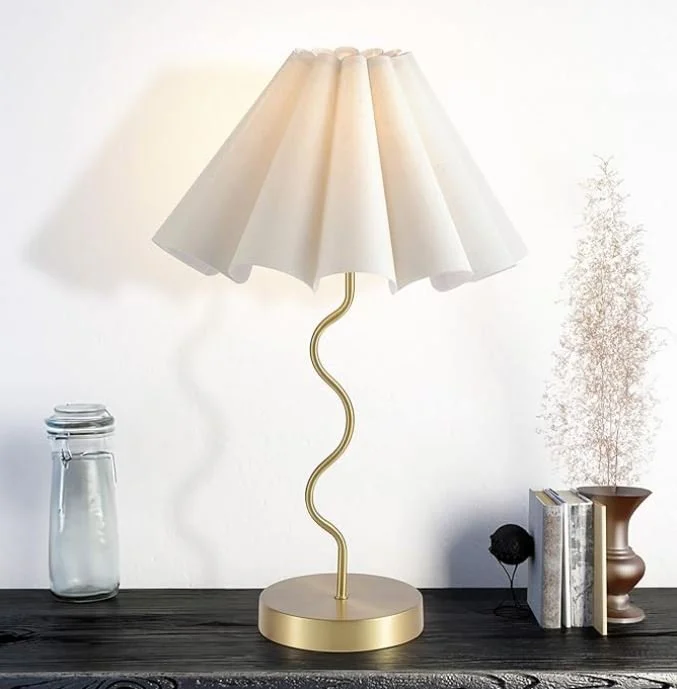 Interior design Inspiration featuring a stylish shoppable decorative table lamp with a gold, wavy stand and a beige pleated lampshade on a dark wooden surface, next to a glass jar, books, and a small potted plant.