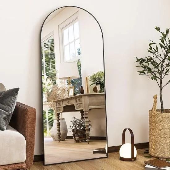 Interior design Inspiration featuring a stylish shoppable full-length mirror with rounded top reflecting a living room area with a side table, lamp, decorative items, and a window with greenery outside, placed against a white wall.