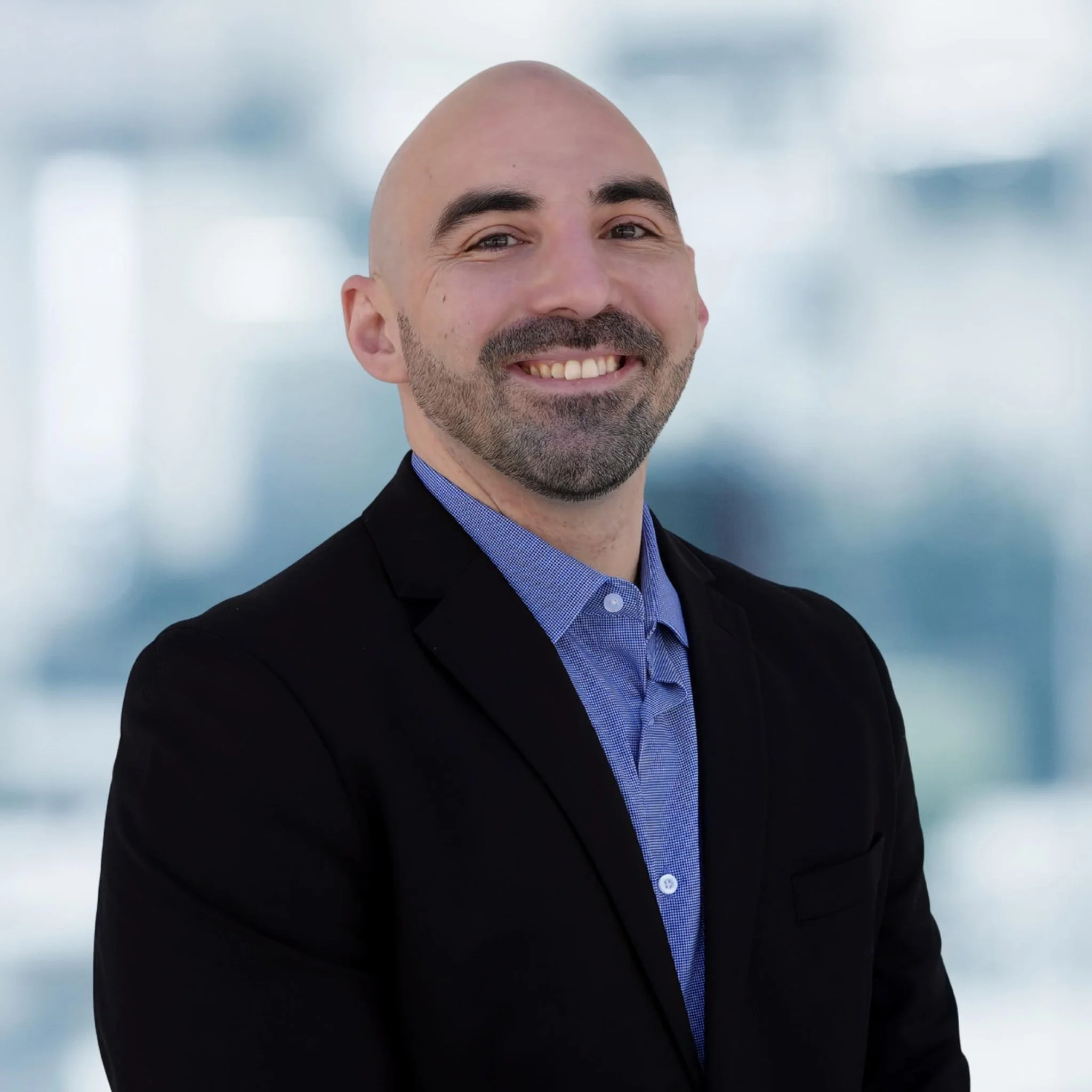 A man with a bald head and beard, wearing a black blazer and a blue shirt, smiling at the camera with a blurry office background.
