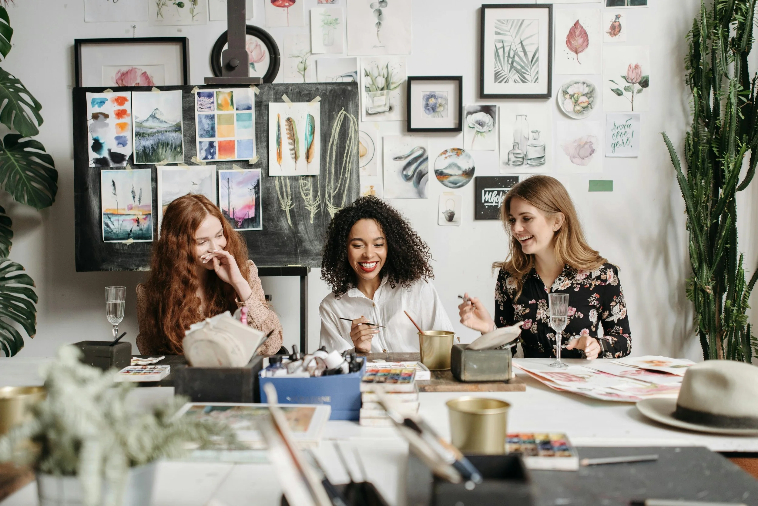 Three women sitting at a table filled with art supplies, laughing and sharing a moment in an art studio decorated with various paintings and botanical illustrations.