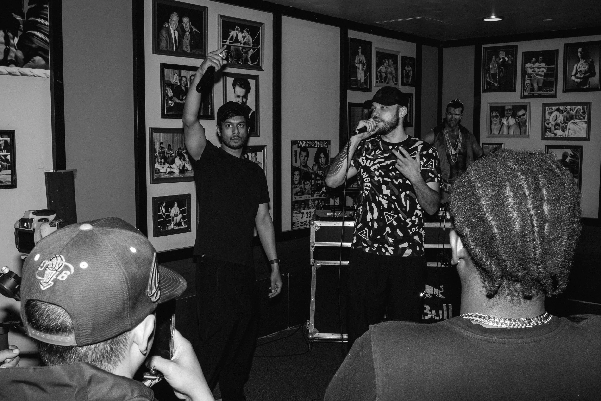 A Black and white photo of HumLo performing on stage at an indoor venue surrounded by photos on the walls. One man holds a microphone and gestures with his hand, while another man stands beside him. Audience members are visible in the foreground.
