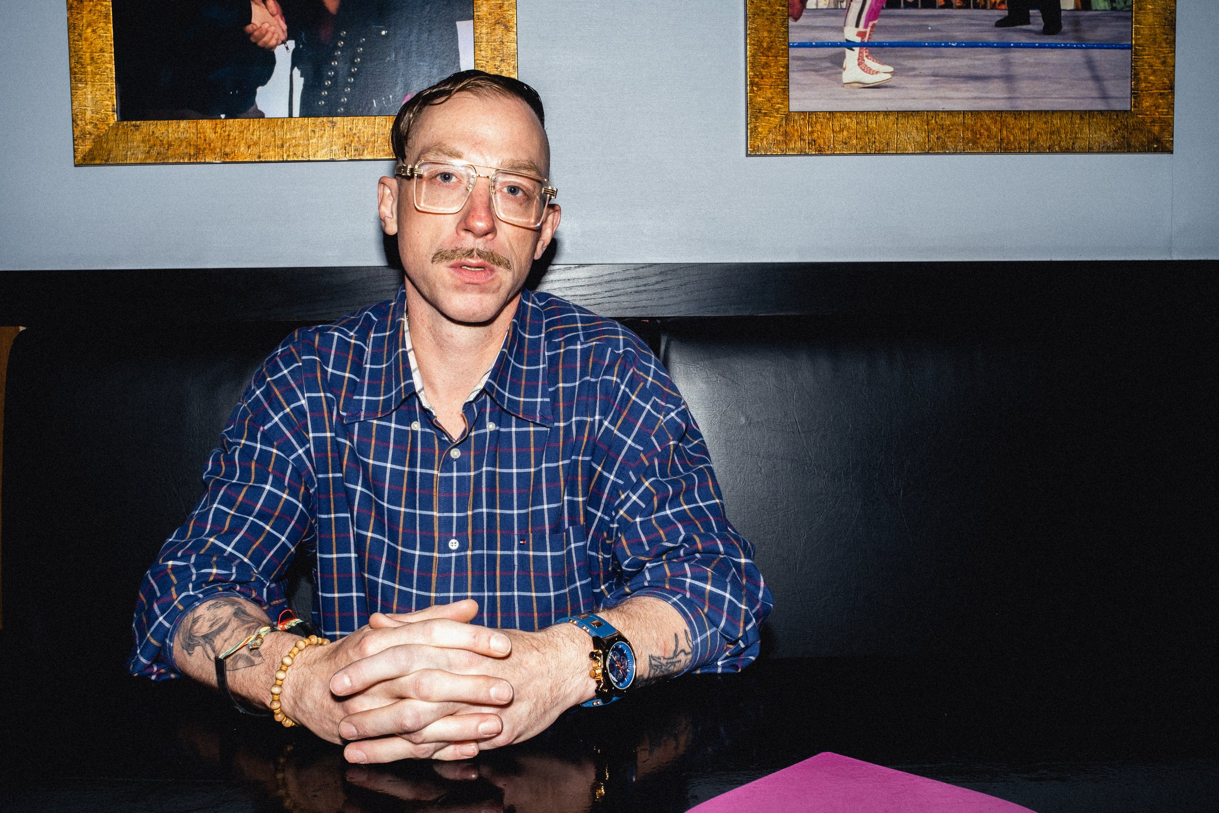 A man with glasses, a mustache, and tattoos sitting at a black table, in front of a gray wall with framed artwork.