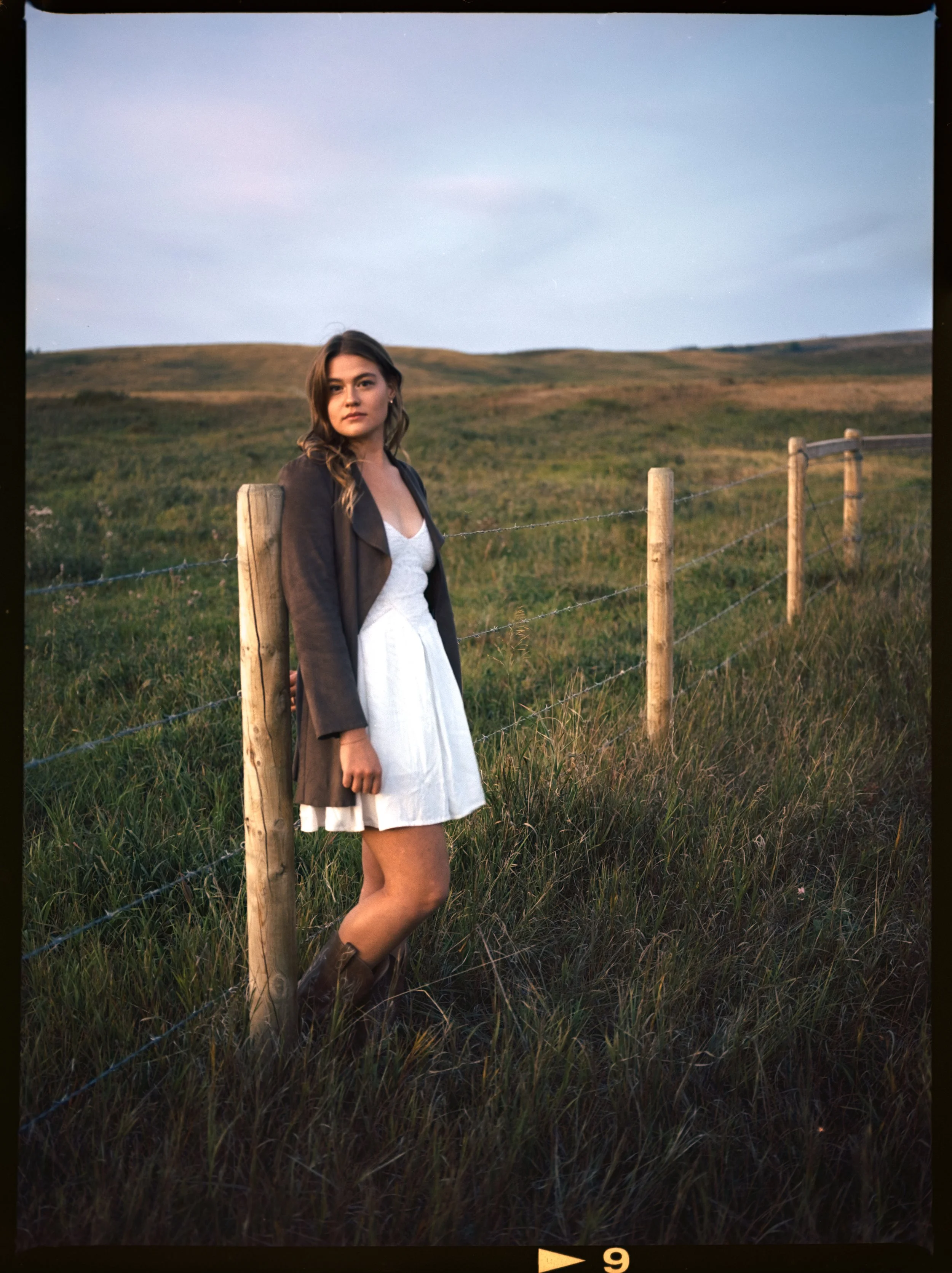 Editorial Portrait: A young woman standing beside a wooden fence in a grassy field during daytime, wearing a brown jacket, white dress, and brown boots. Haskayne Park outside of Calgary