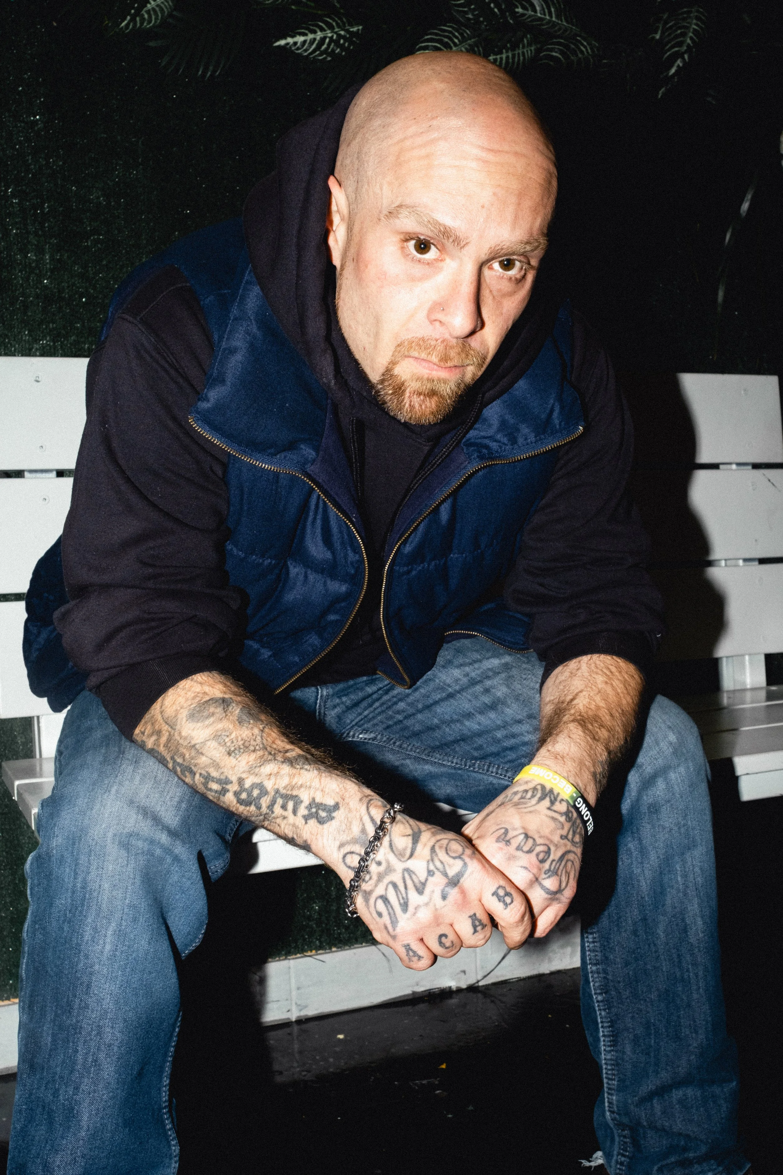 Artist Portrait: "RYE'N WiLD" A man Sitting on a bench leaning forward wearing a Vest and Jeans.