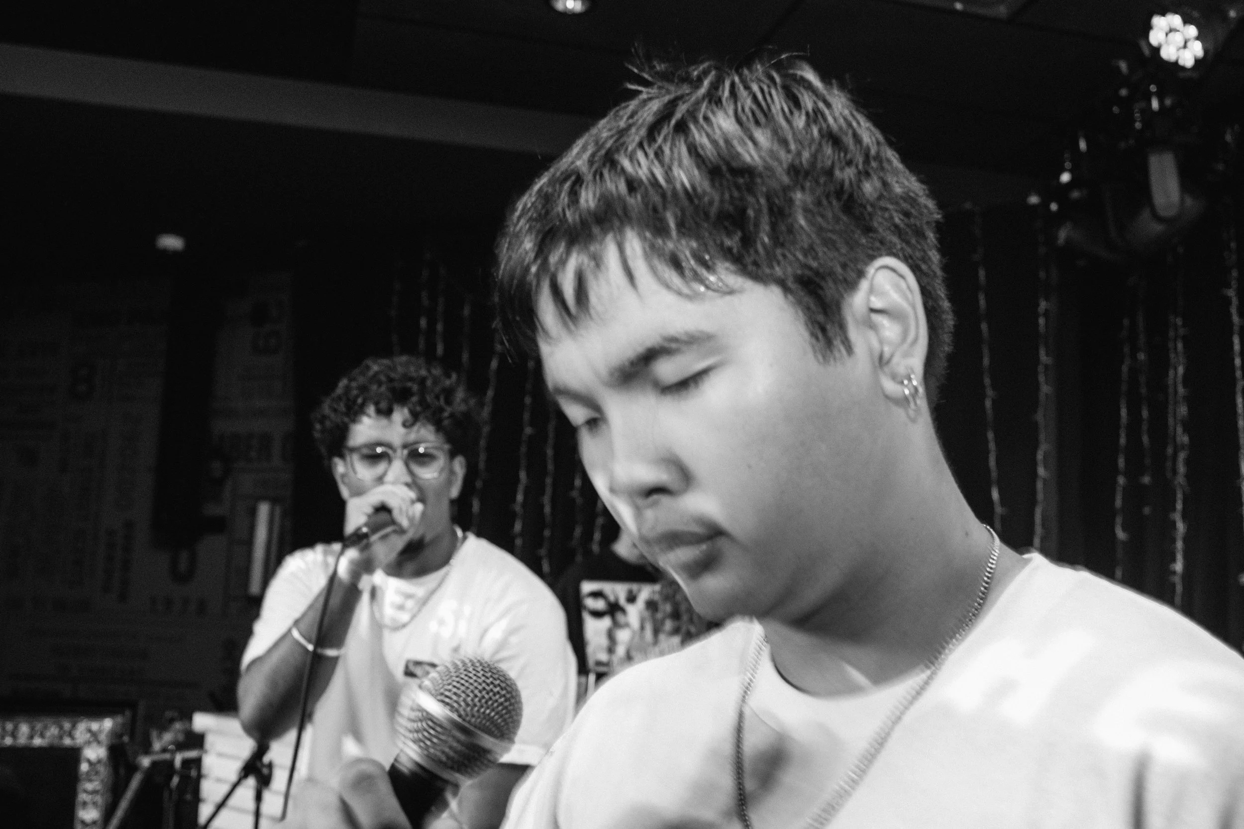 Typical and Jaywo performing music, one holding a microphone and the other with eyes closed, in a setting with dark curtains and stage lighting.