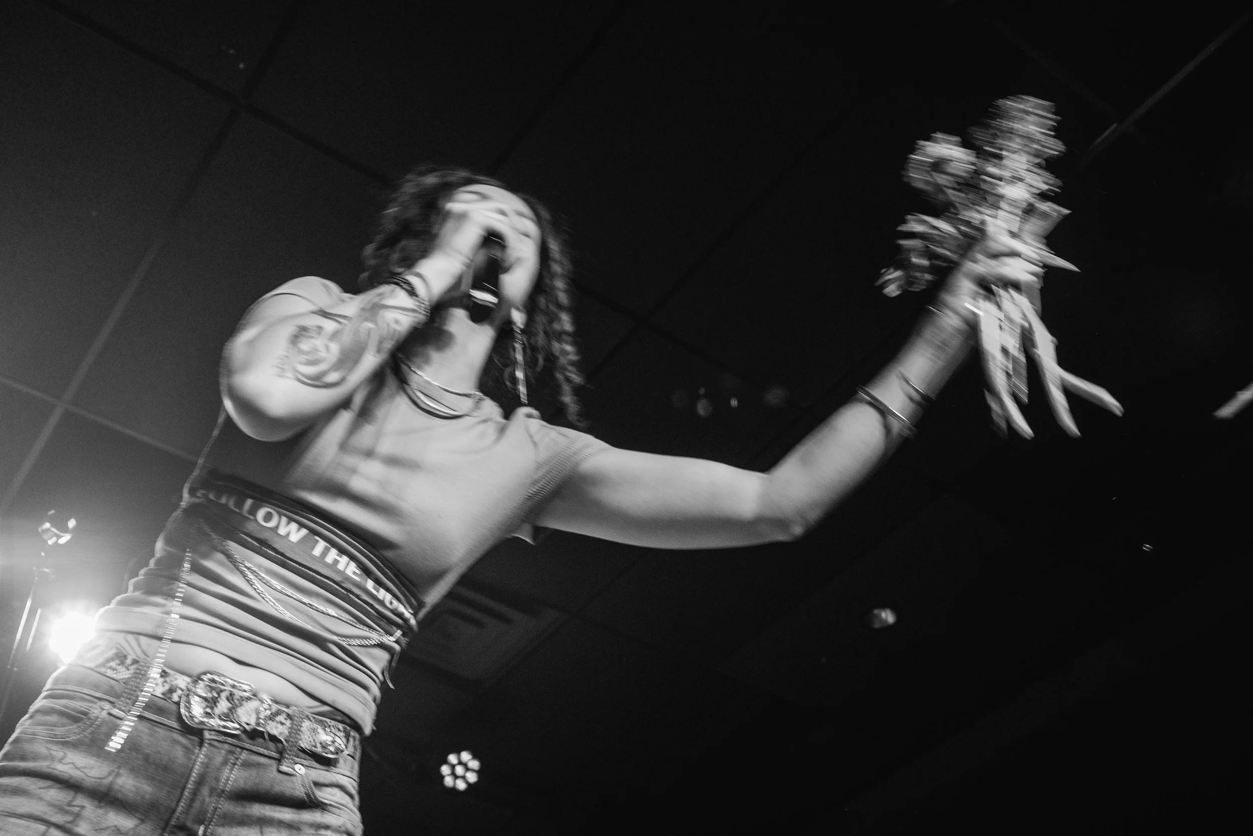 Amarie performing, wearing a t-shirt and jeans, is on stage holding a microphone close to her mouth with her left hand and a bunch of money in her right hand. The image is in black and white.