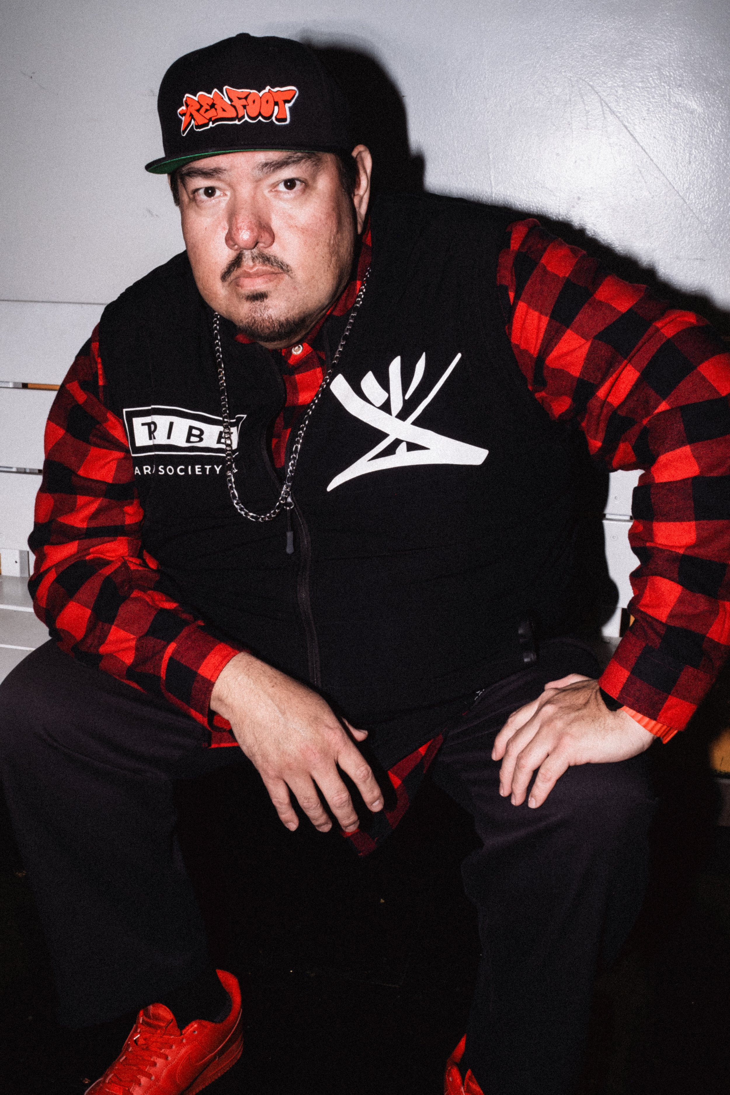 Artist Portrait "Redfoot". A man sitting with his arm on his leg, black hat, red flannel and black vest.
