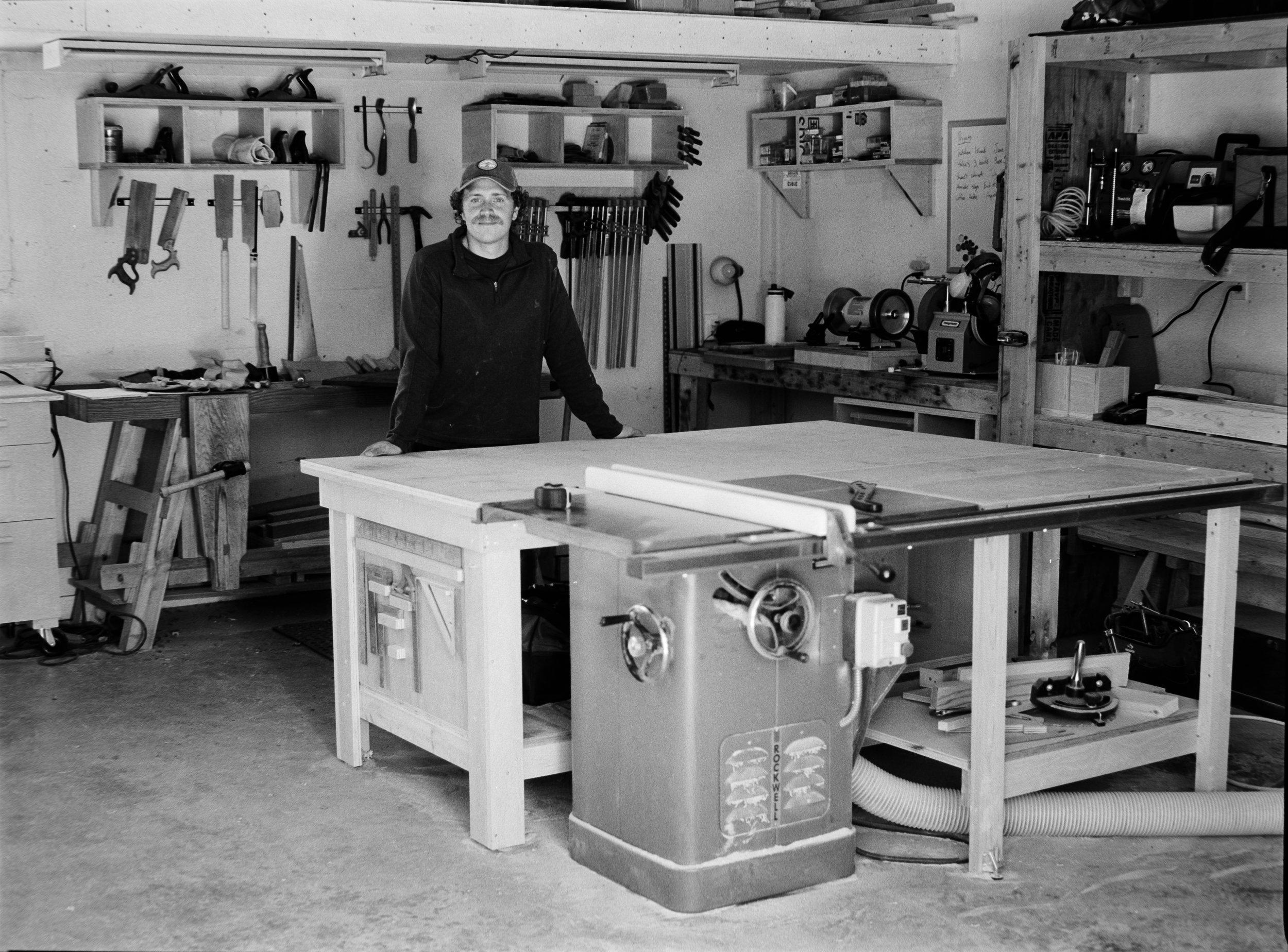 A person standing in a woodworking workshop surrounded by various tools and equipment, including a large table saw in the foreground. editorial photographer calgary