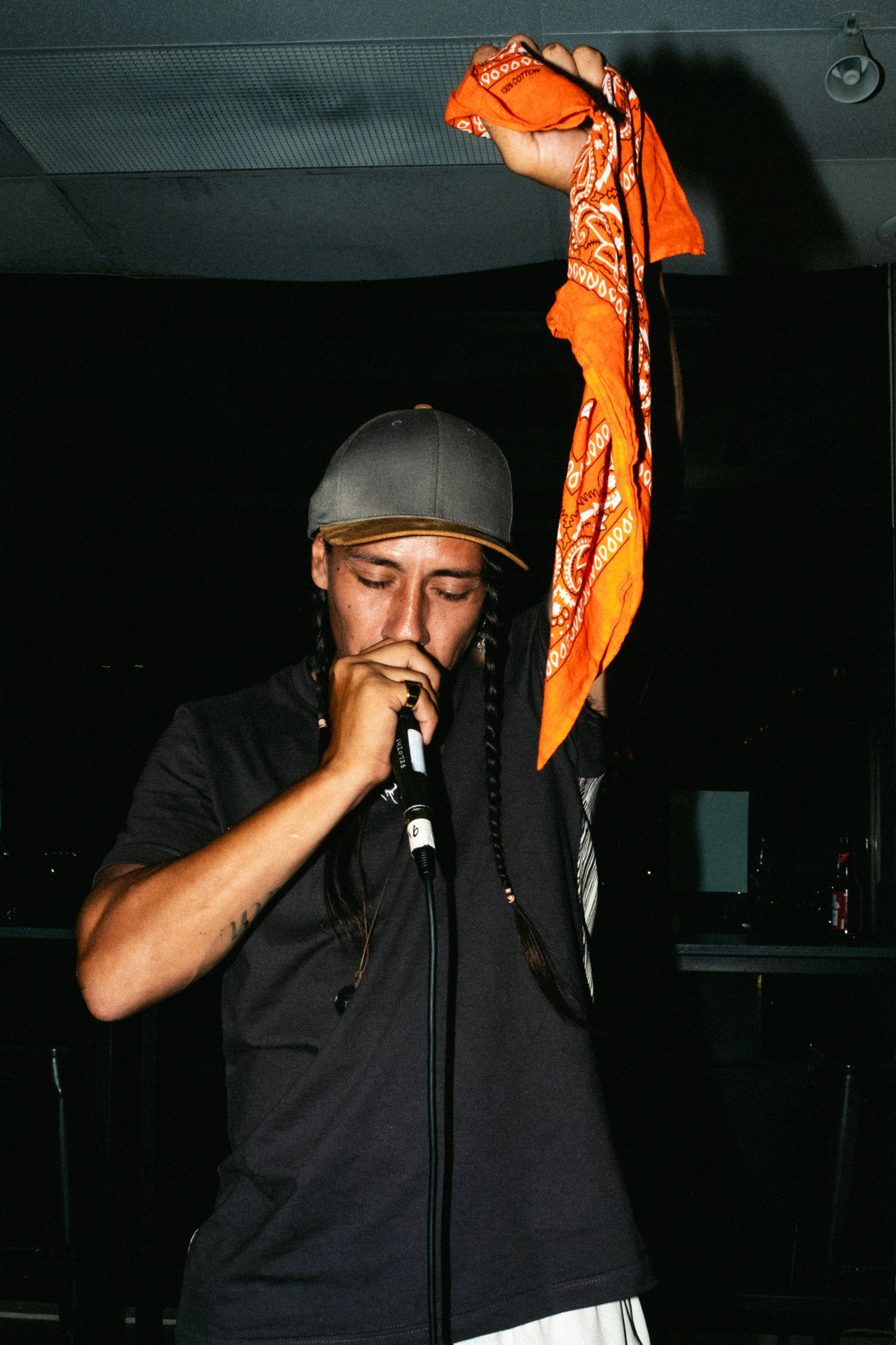 Tecun B'alam on stage with a microphone in one hand raised holding an orange bandana, wearing a black shirt and a gray baseball cap, with braided hair, in a dark indoor setting.