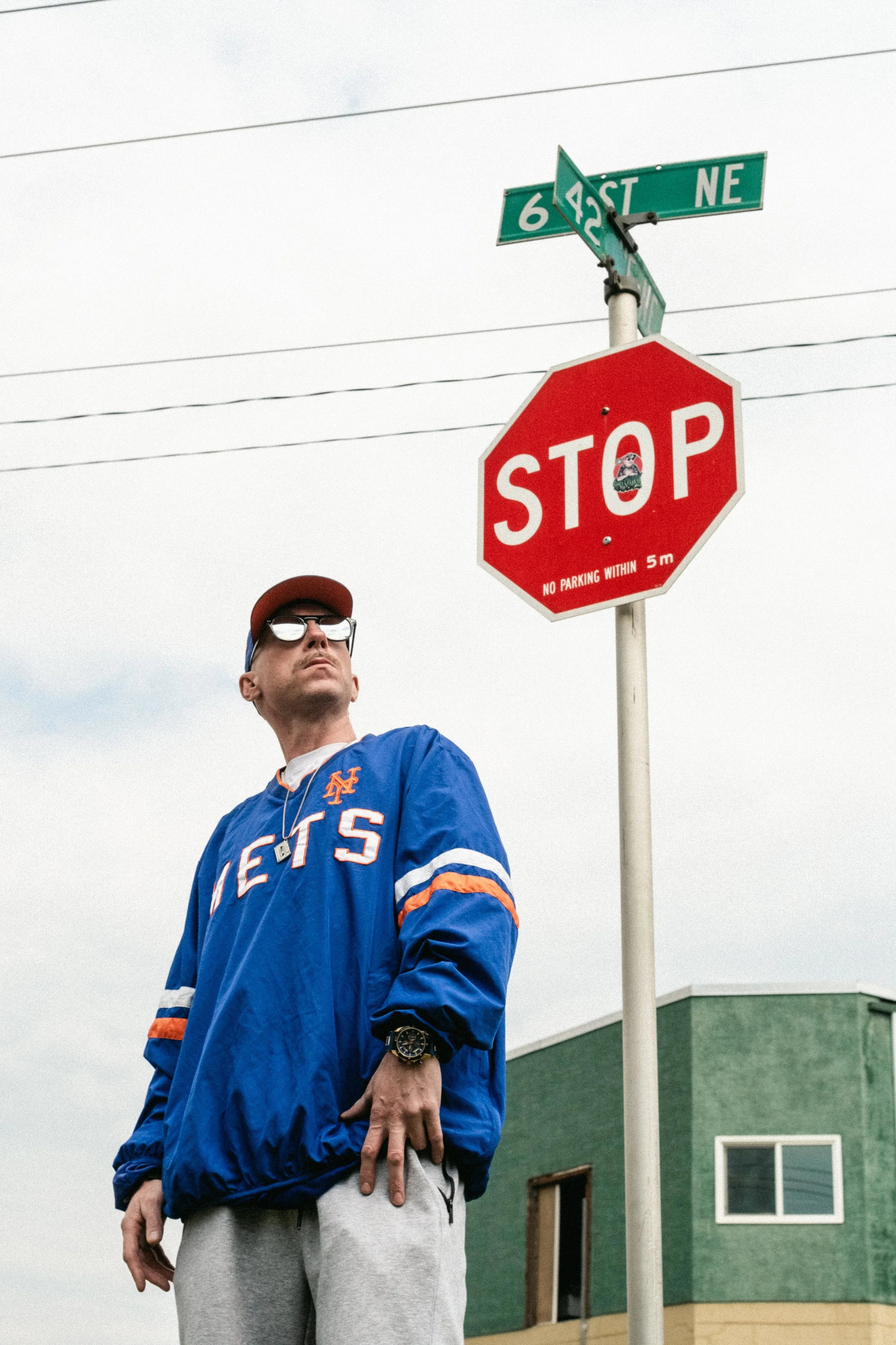 Artist Portrait: "THE ViNZ". Wearing a New York Mets jacket, sunglasses, and a cap, standing under a stop sign at an intersection with street signs with a green building in the background.