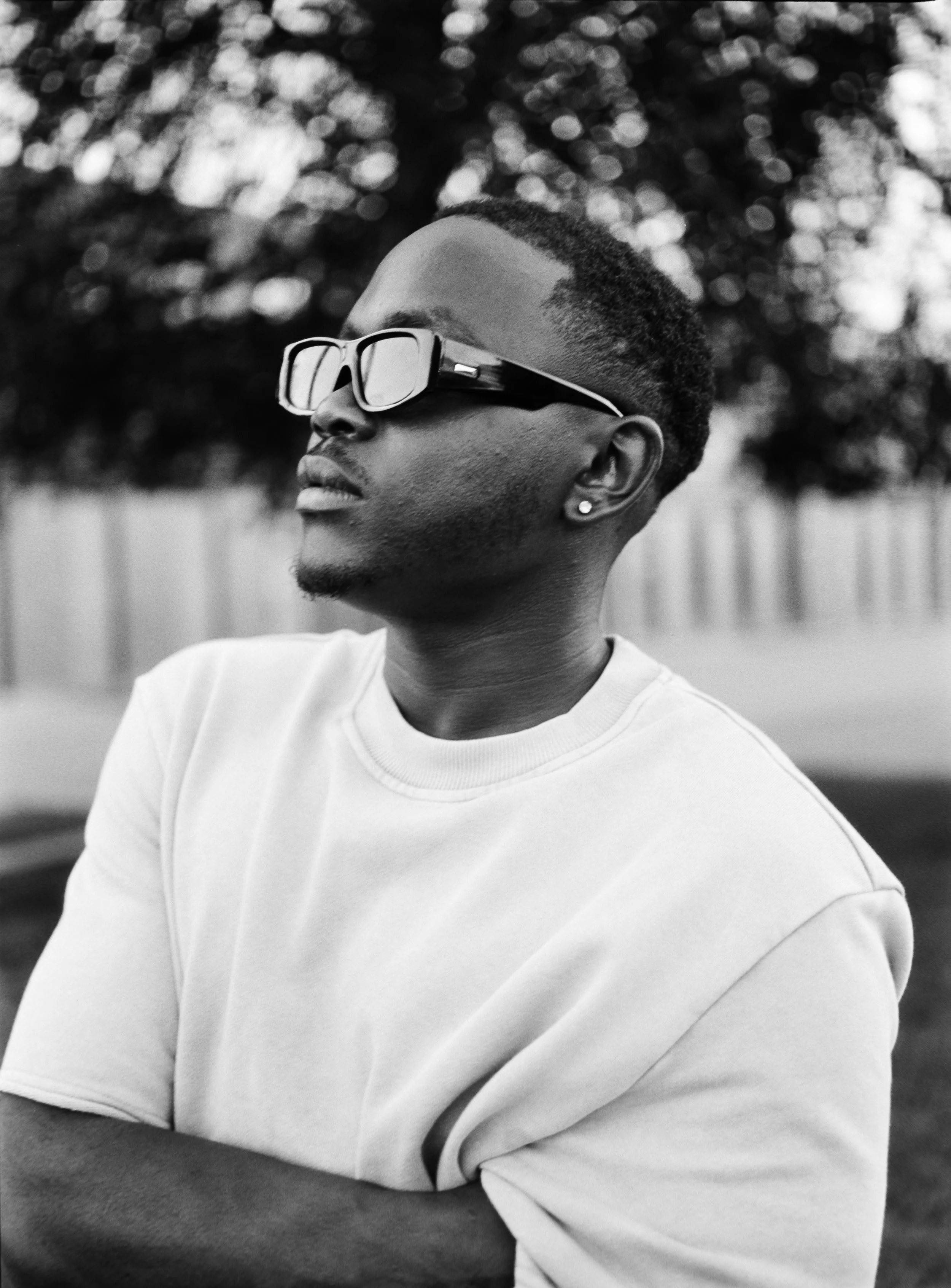 Artist Portrait: "Types". Black and white photo of a young man with short hair, wearing sunglasses and a white t-shirt, with arms crossed, standing outdoors near trees and a fence.