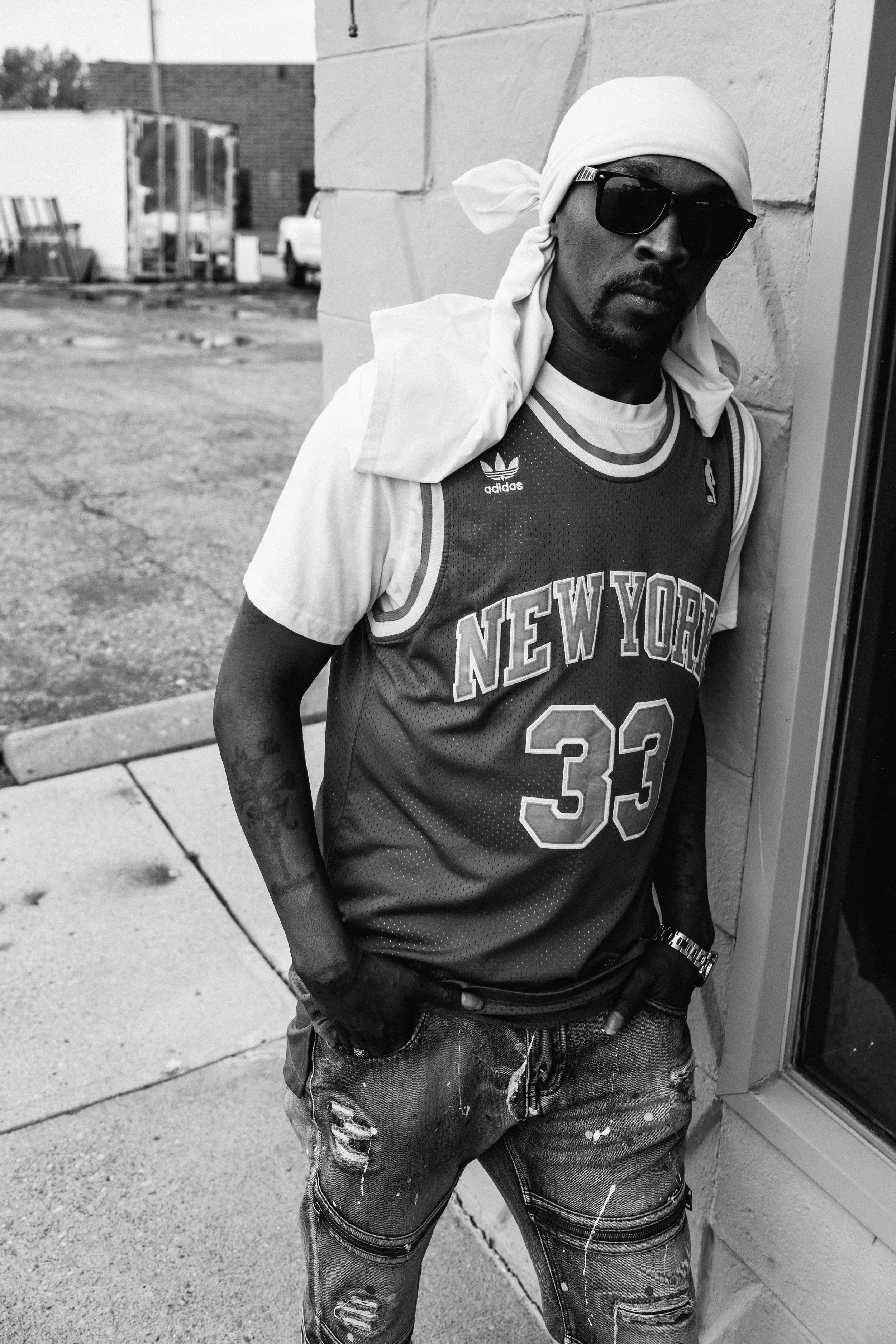 Artist Portrait: "Risky G". A man wearing a New York Knicks basketball jersey with the number 33, sporting sunglasses, a white bandana or headscarf, and distressed jeans, standing outside against a brick wall with his hands in his pockets.
