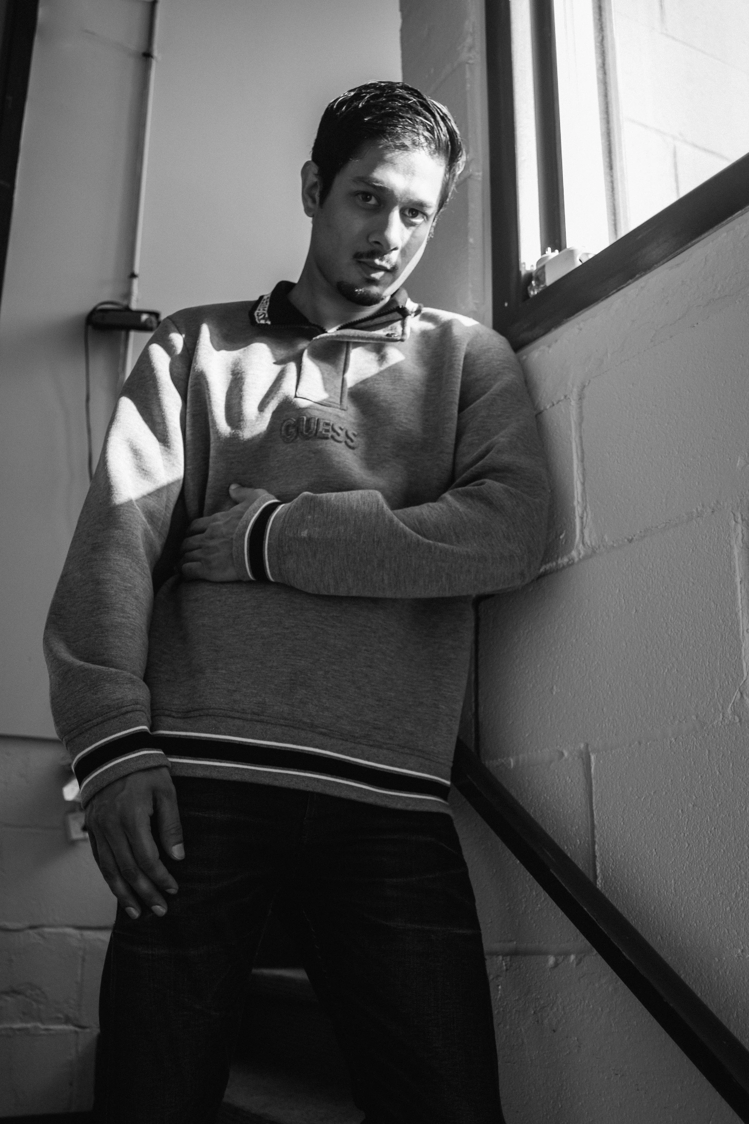 Artist Portrait: "HumLo FrqNtc". A young man with dark hair and a goatee, wearing a grey GUESS sweatshirt, standing against a brick and wall corner near a window, looking at the camera with a serious expression.