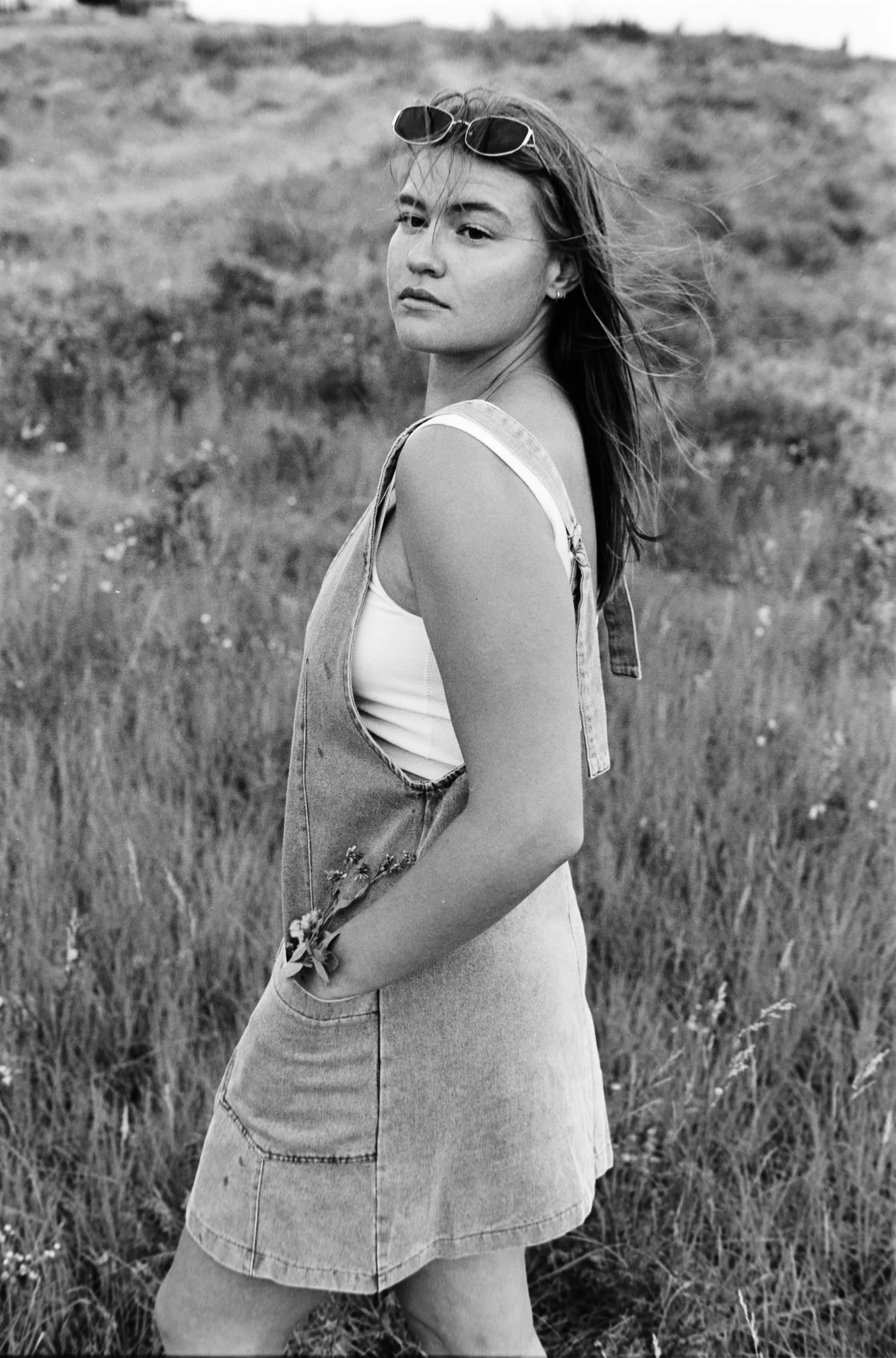 Black-and-white photo of a woman standing in a grassy field, wearing a sleeveless top and denim dress with sunglasses on her head, looking over her shoulder.