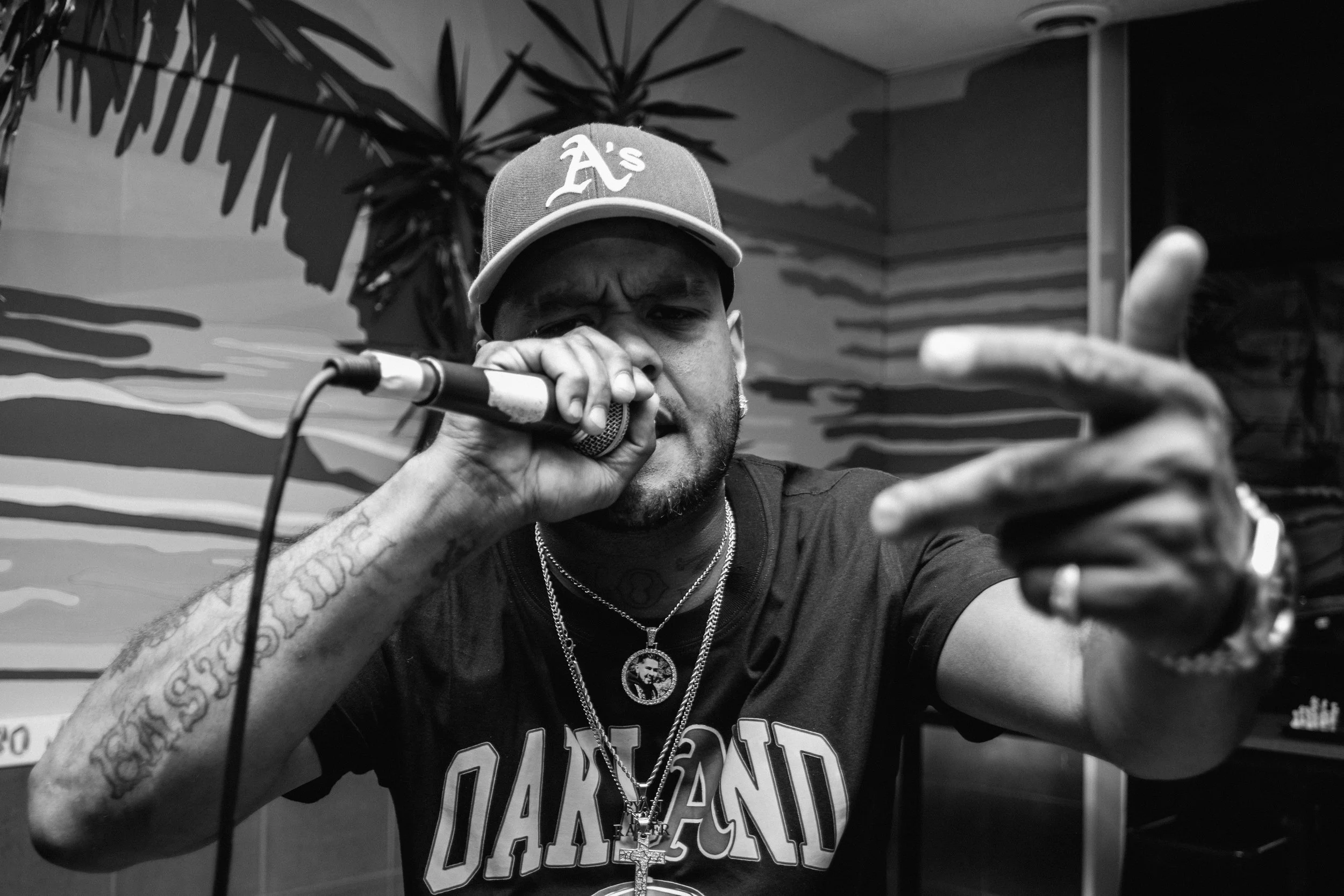 Ryan The Rapper wearing a Oakland baseball cap singing into a microphone with a serious expression, pointing forward with his other hand, in a room with mural art and a tropical plant in the background.