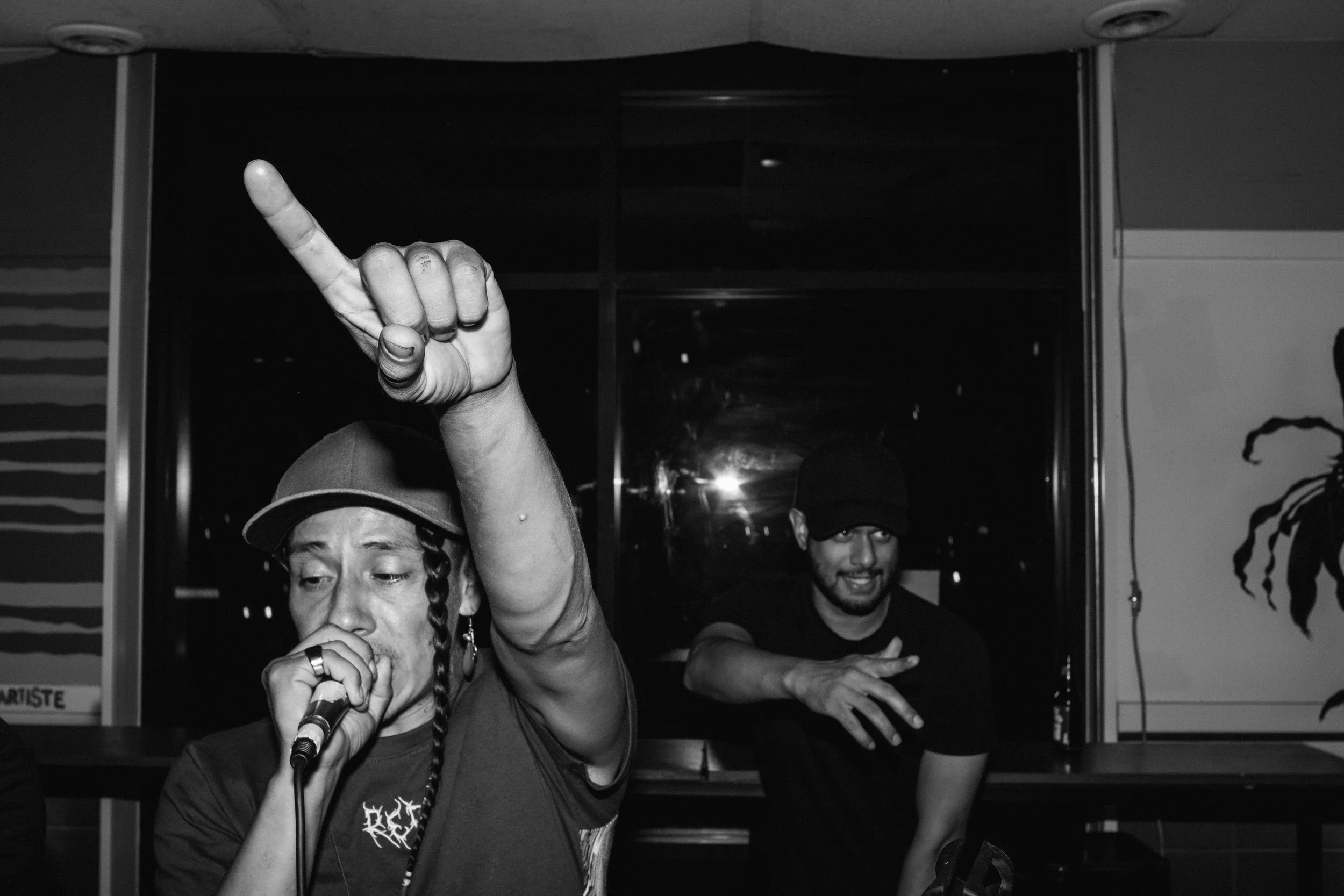 Tecun B'alam Performing, holding a microphone, singing or speaking with her eyes closed, and raising her hand with her index finger pointing upwards, while a man in a black cap and t-shirt stands behind her, smiling and extending his hand.