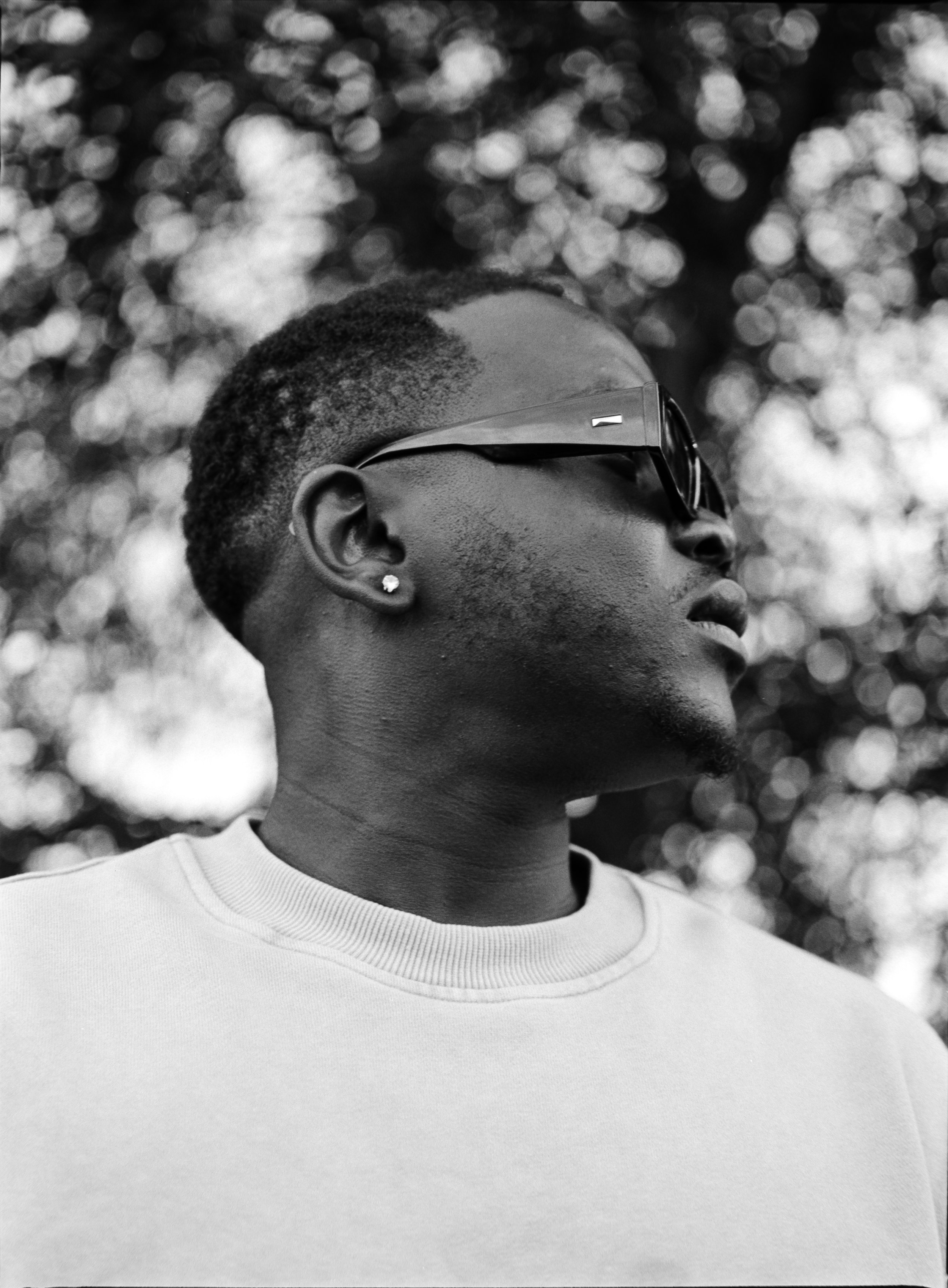 Artist Portrait: "Types". A young person with short hair, wearing sunglasses and a stud earring, looking to the right, with a blurry background of trees.