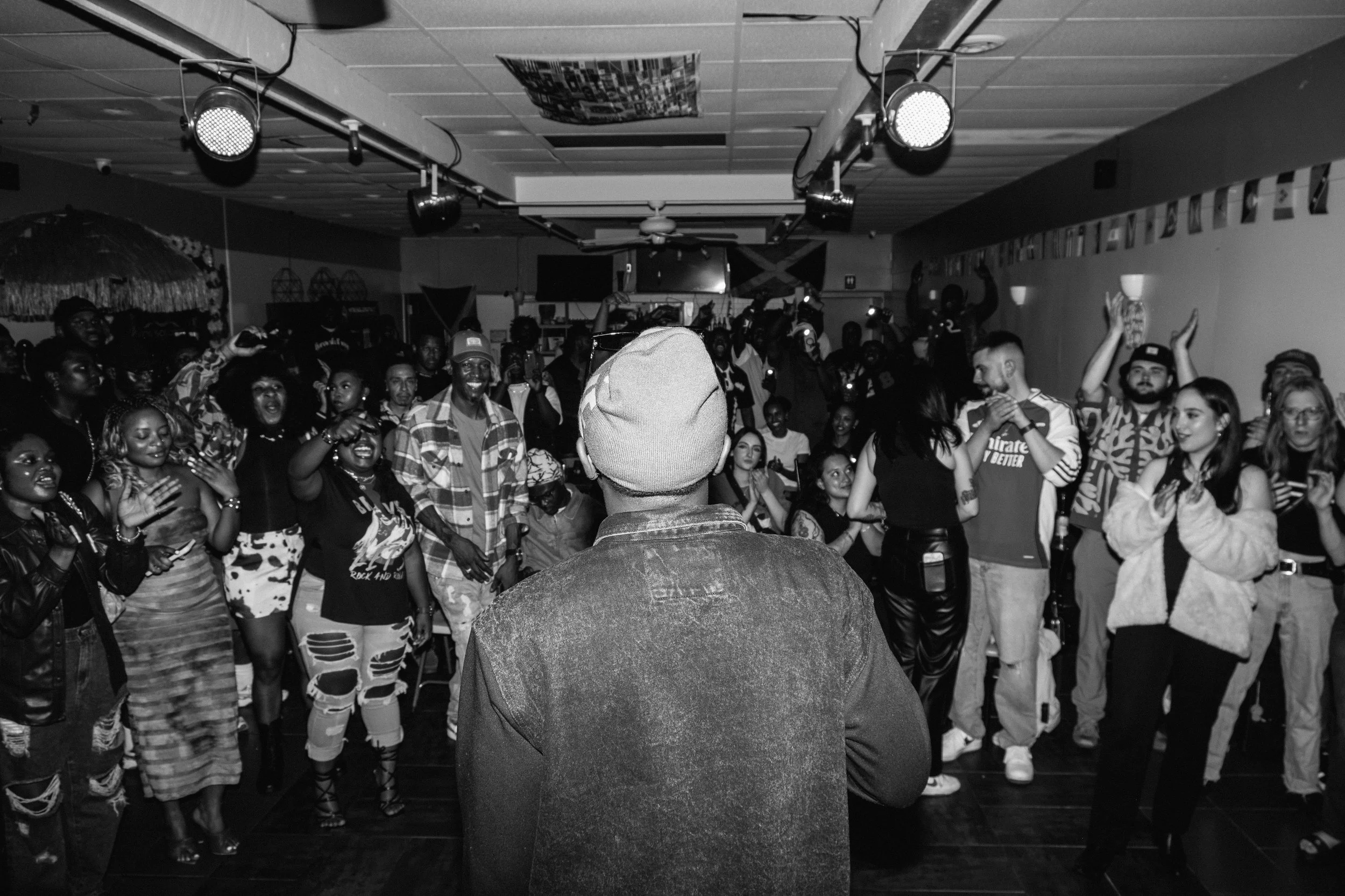 Types On stage. A black and white photo of a crowded indoor party, seen from behind a person wearing a hat and denim jacket, facing a lively group of people dancing and celebrating.