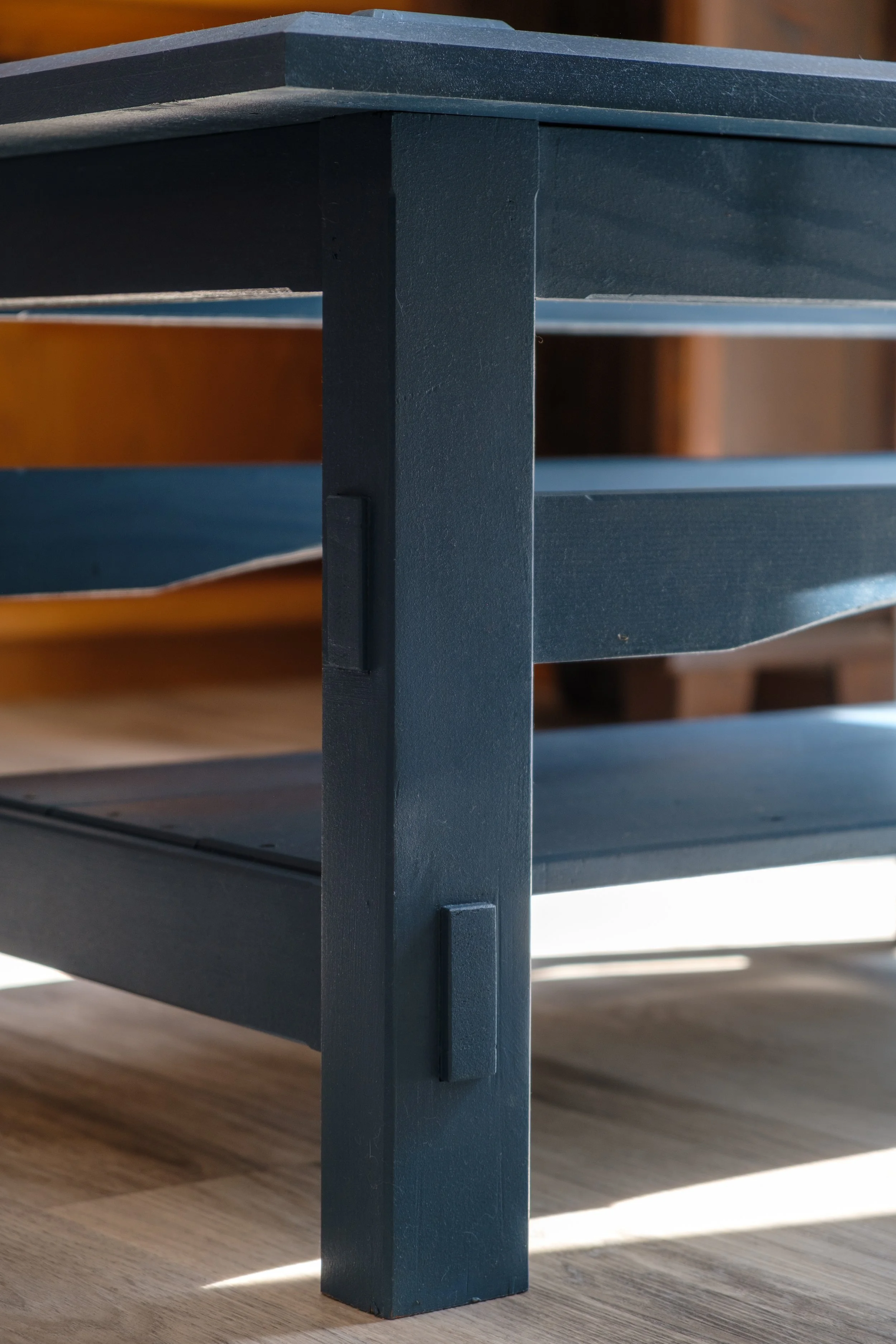 Product Photoshoot for Klein Woodworks. Close-up of a blue wooden furniture piece, showing the corner with a vertical leg and horizontal supports, set on a wooden floor with sunlight casting shadows.