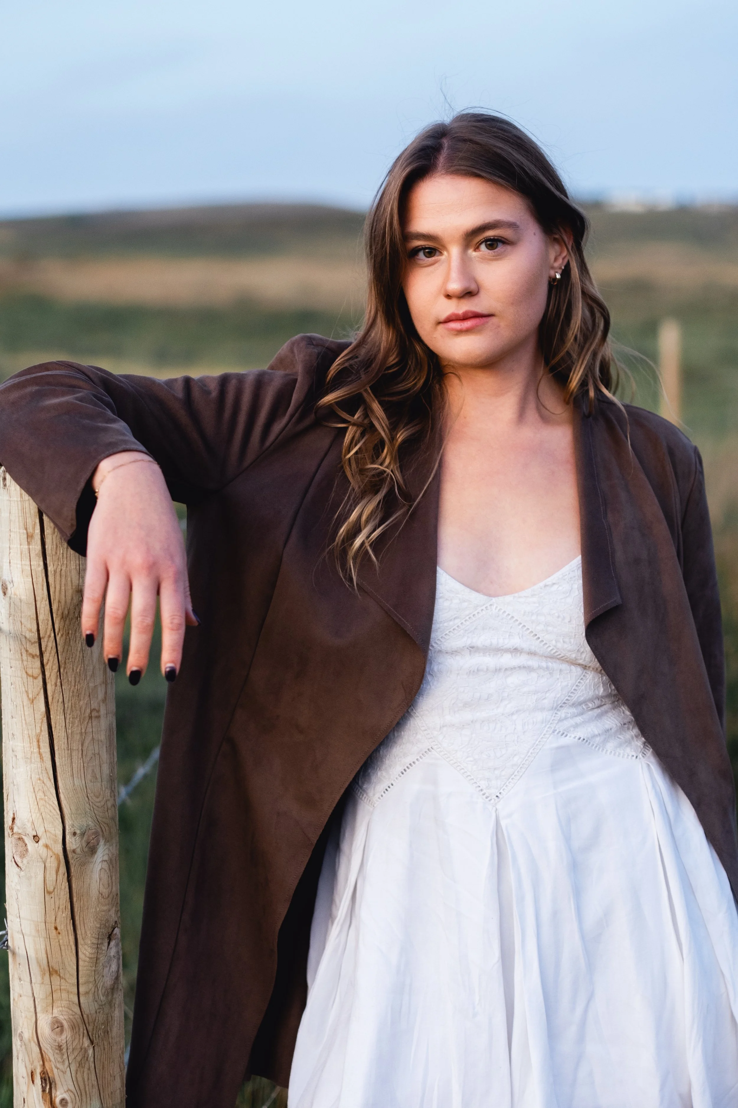 Fashion Photoshoot Calgary. A young woman with long, wavy brown hair leaning on a wooden fence post outdoors. She is wearing a brown jacket over a white dress, slightly standing in a field with rolling hills in the background during daylight.