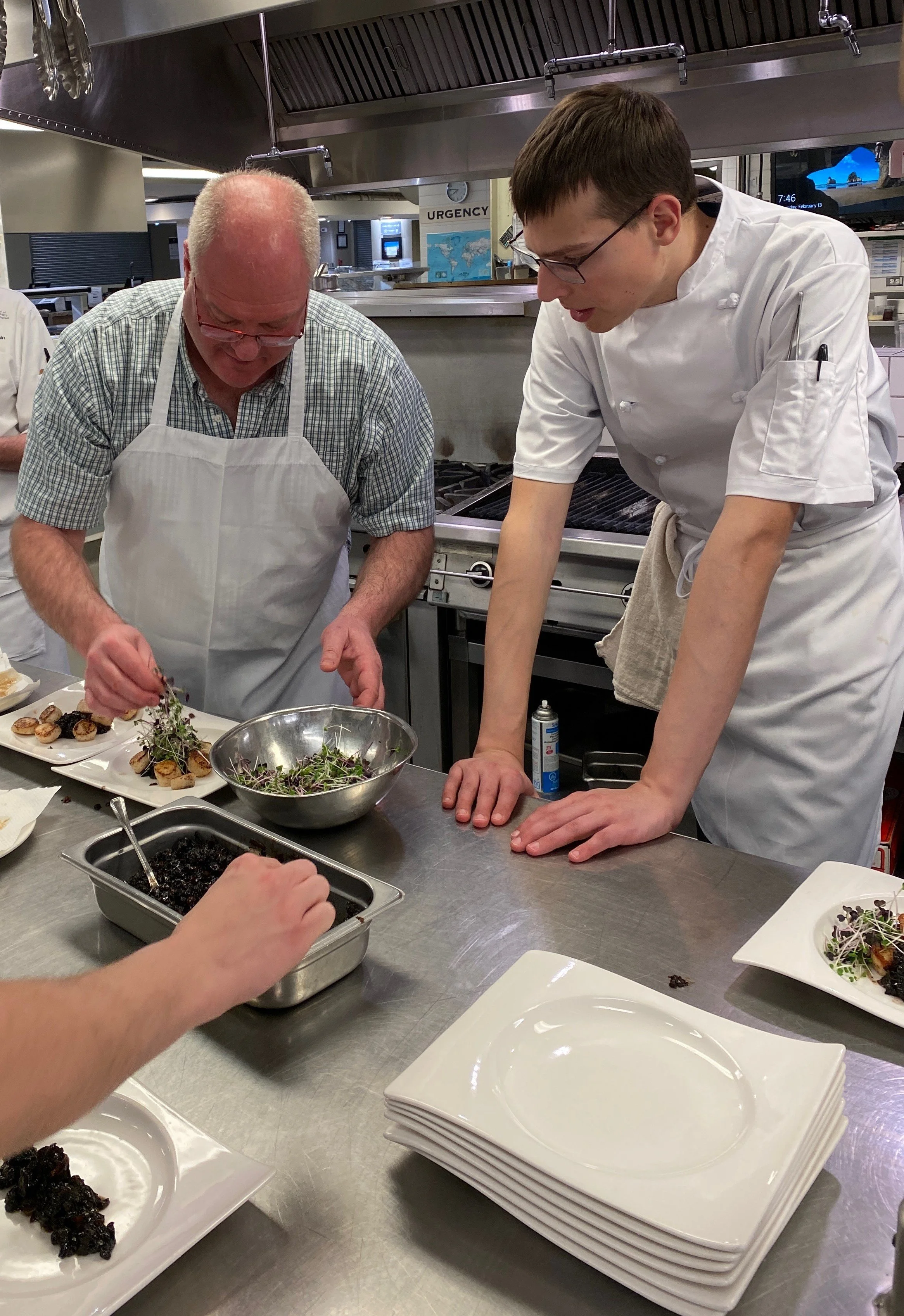 Two chefs are preparing dishes in a commercial kitchen with plates and ingredients on a stainless steel counter.