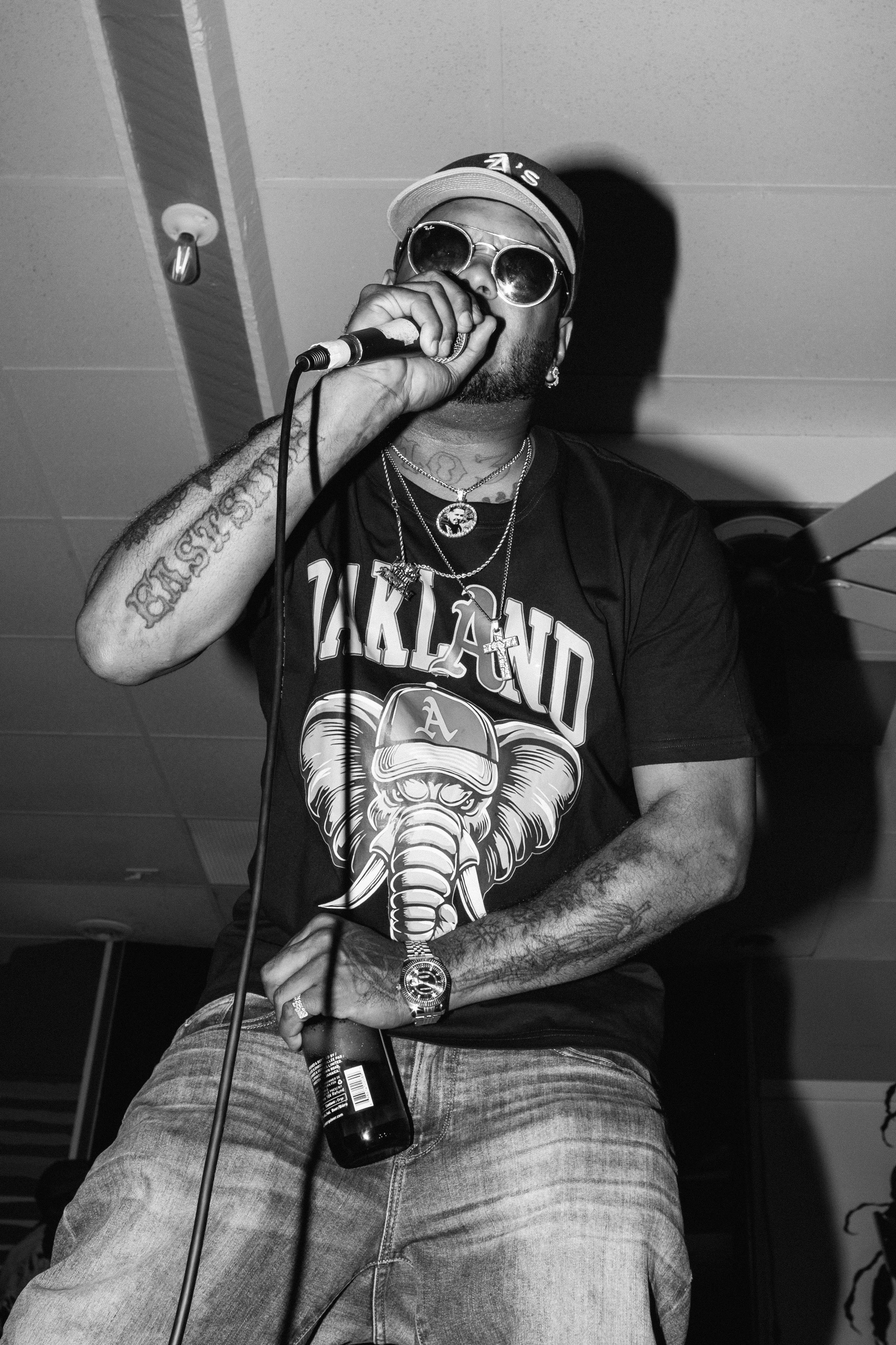 Ryan The Rapper wearing sunglasses, a cap, and jewelry is holding a microphone and singing or speaking, with a bottle of red stripe beer in his hand.