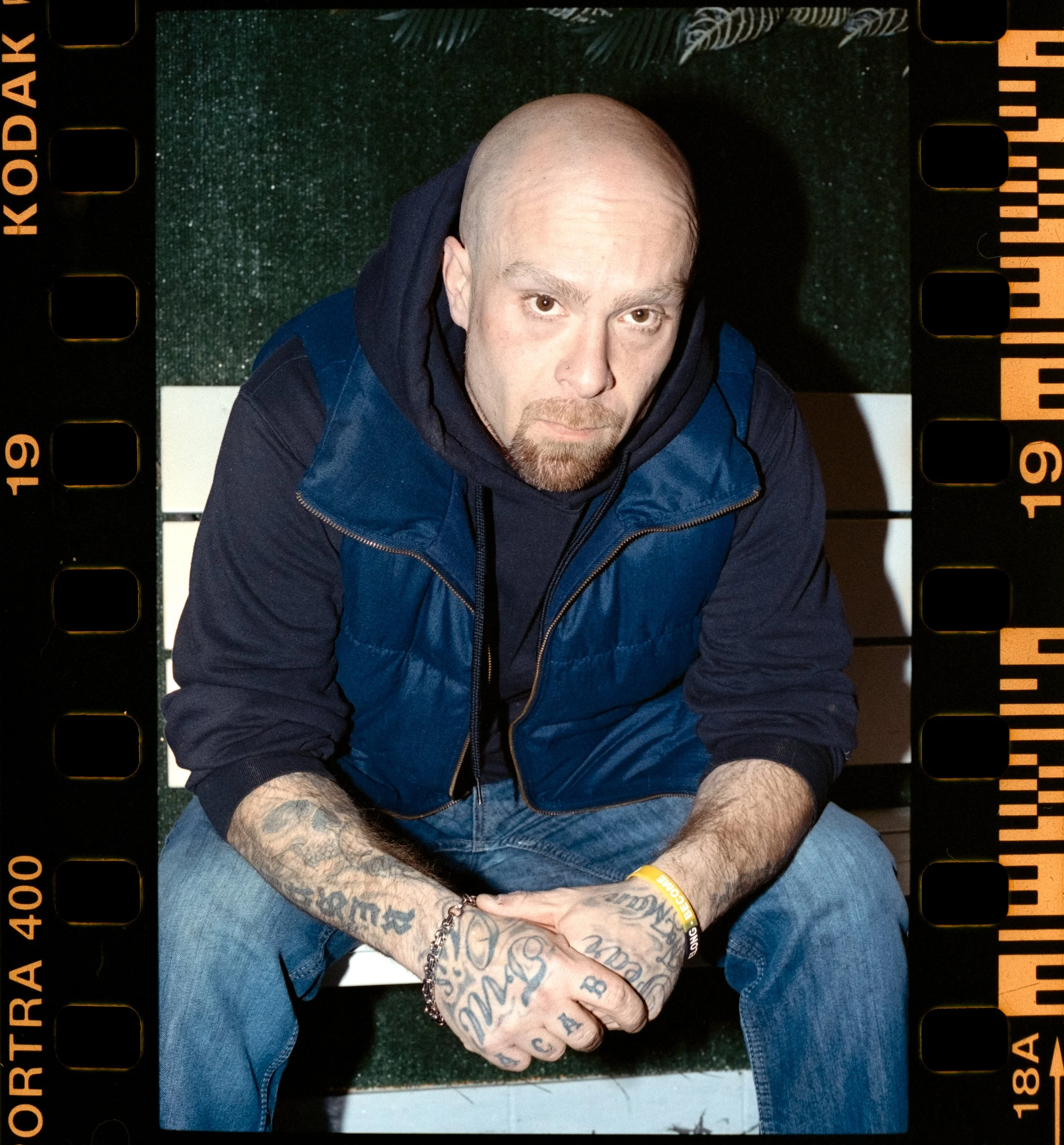 Artist Portrait: "RYE'N WiLD" A man Sitting on a bench leaning forward wearing a Vest and Jeans.