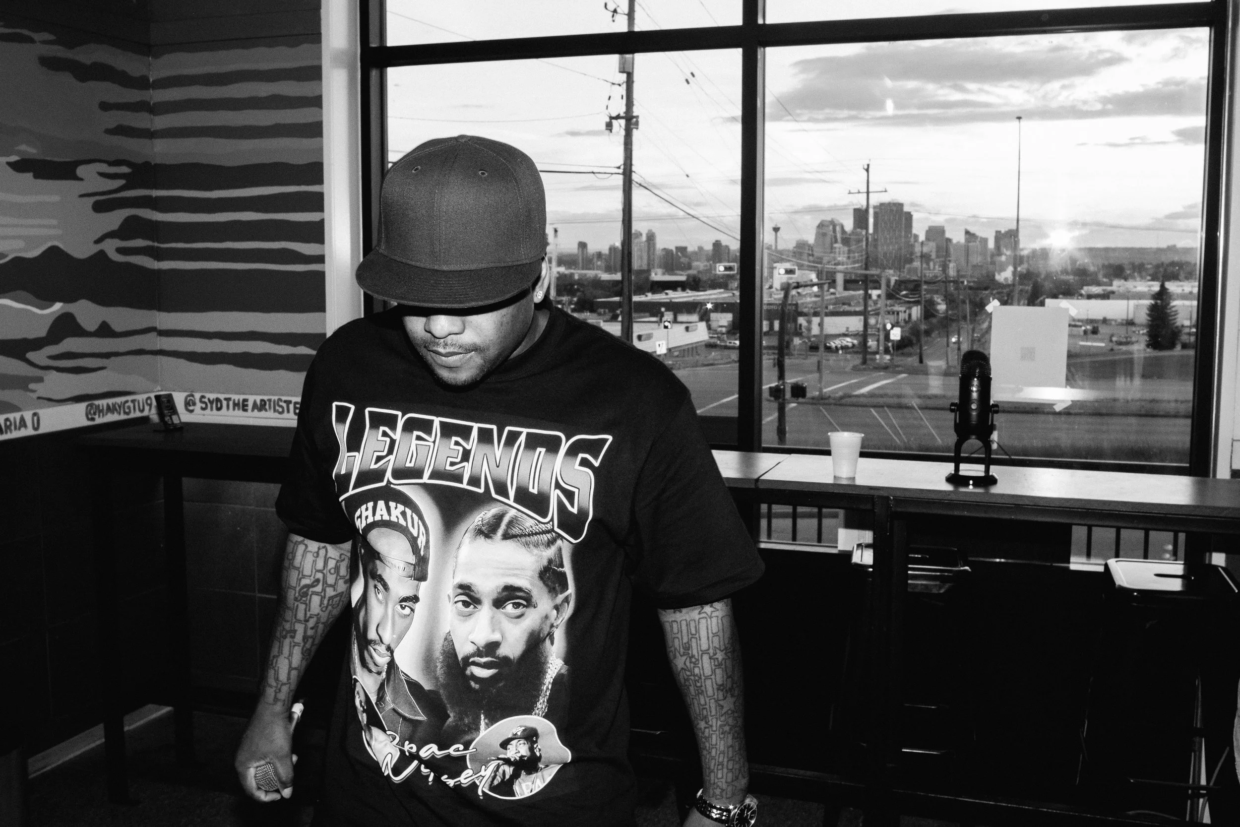 Selloo on stage wearing a black hat and a "Legends" t-shirt with portraits of Tupac and another rapper, standing indoors with large windows showing a city skyline and streets outside.