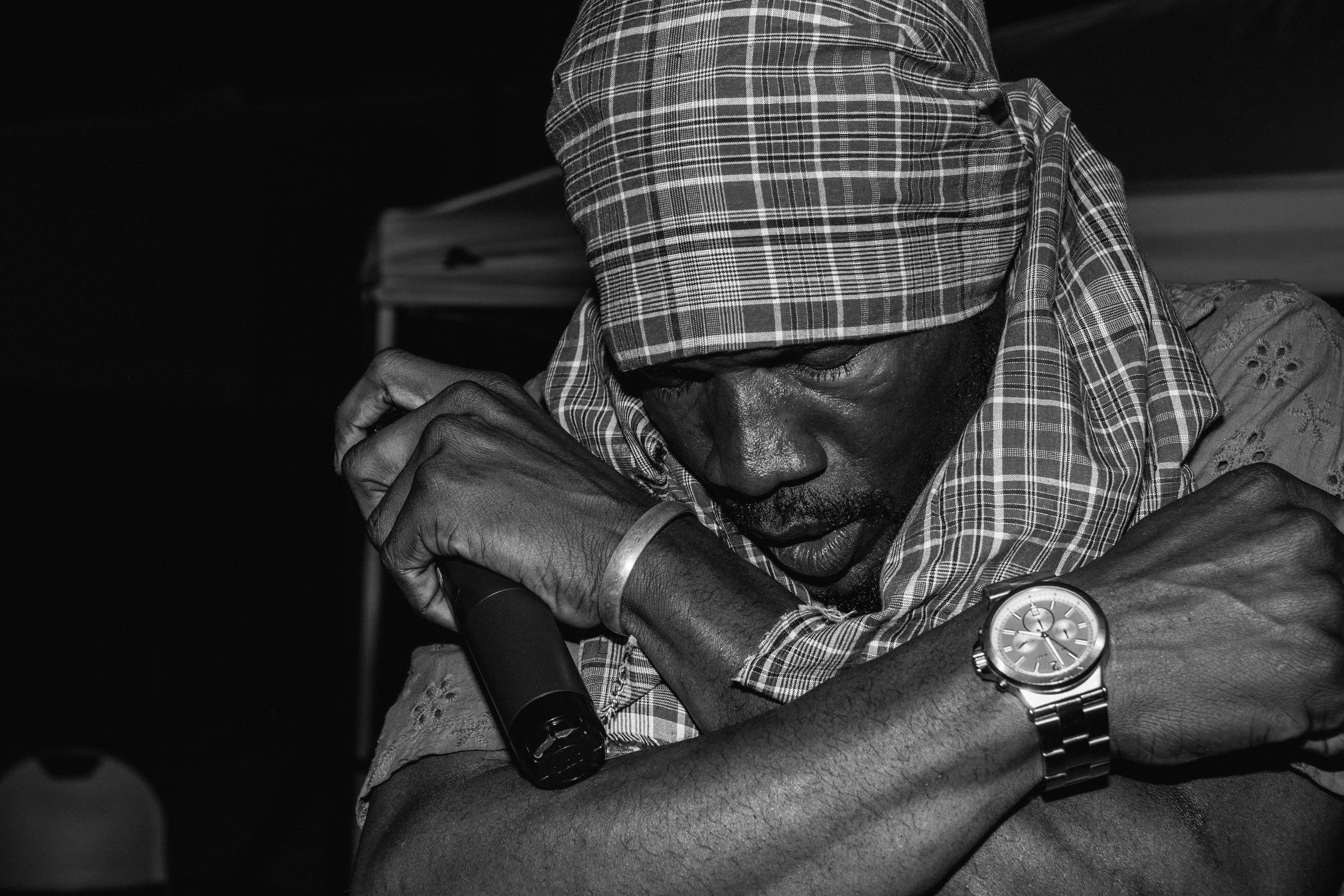 5ive13 on stage in a headscarf and patterned shirt, wearing a wristwatch and bracelet, is wrapping his arms around a microphone with his eyes closed in a moment of emotion.