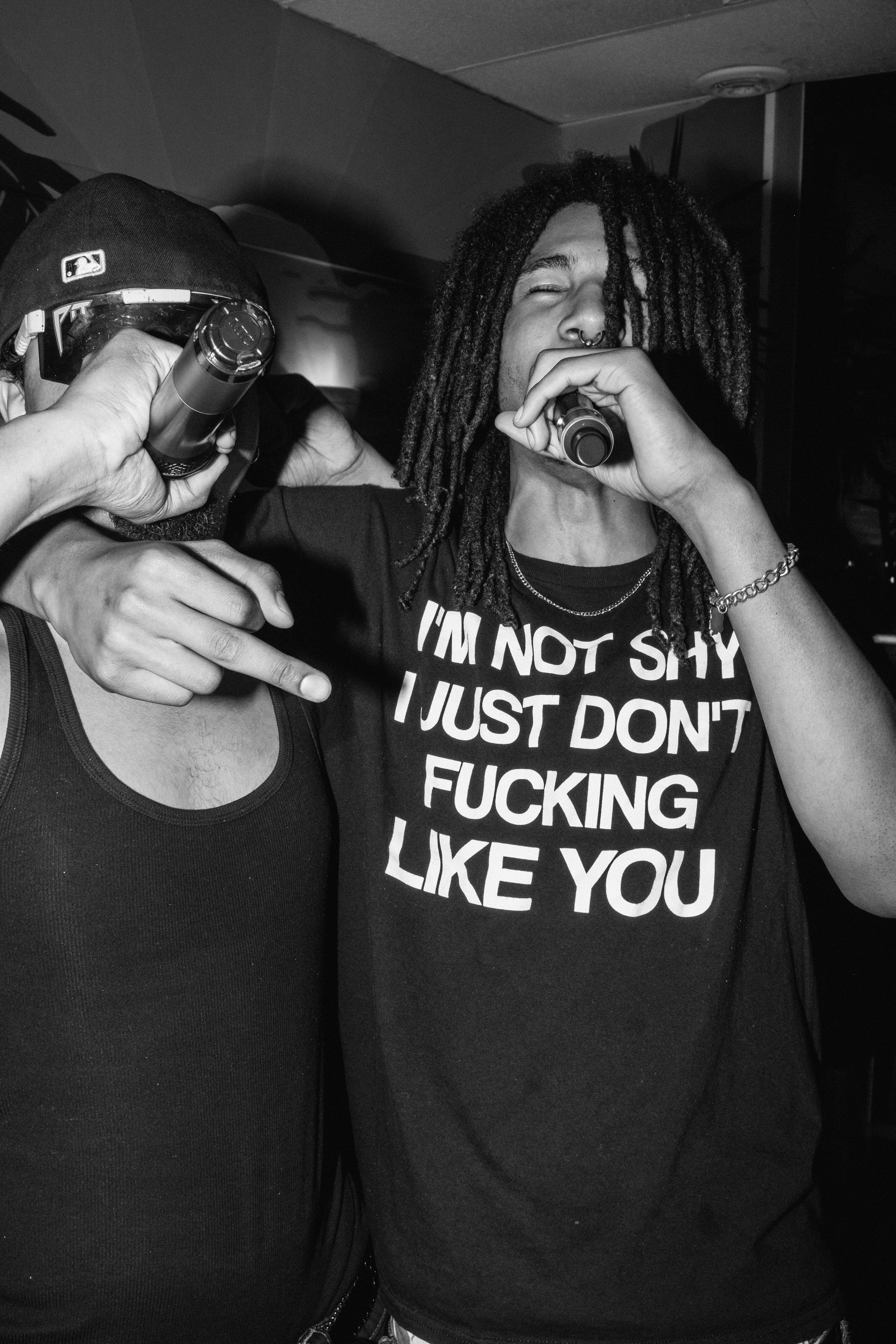 Two young men in a black-and-white photo, one with dreadlocks and the other wearing a cap, are holding microphones. The man with dreadlocks wears a T-shirt that says, 'I'M NOT SHY I JUST DON'T FUCKING LIKE YOU,' and is gesturing with his hand.