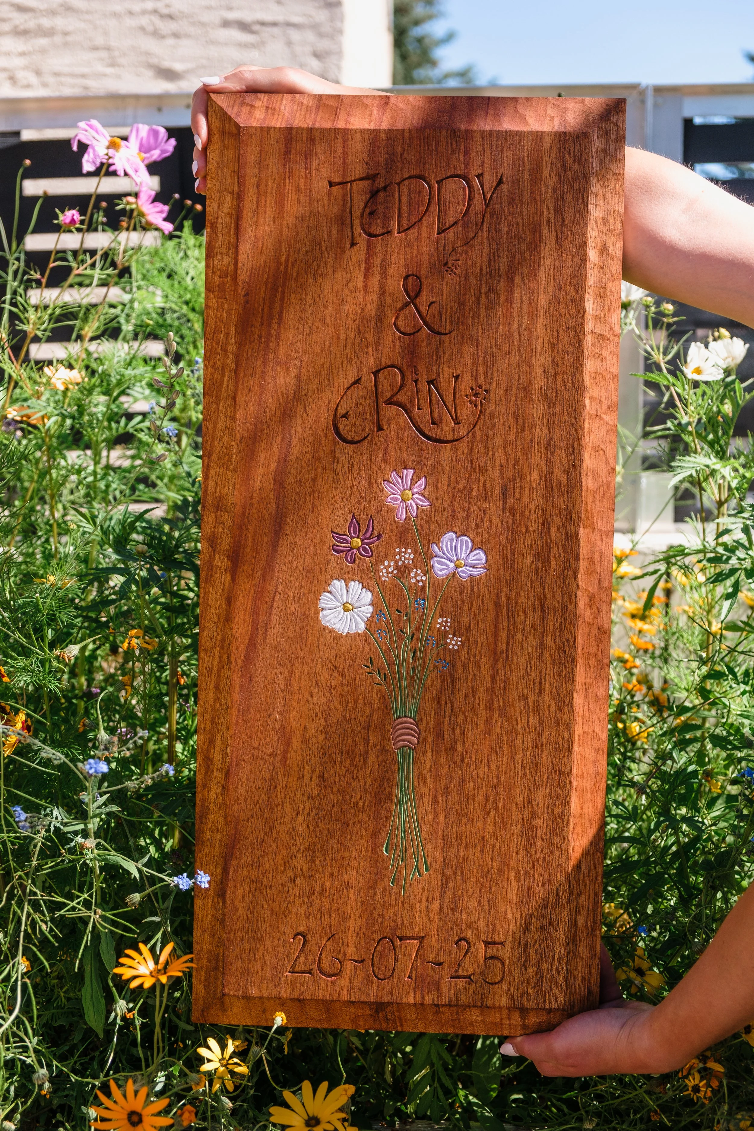 A wooden plaque with engraved text reading 'Teddy & Rina, 26-07-25', decorated with a painted bouquet of flowers, being held by a person among flowers and plants outdoors.