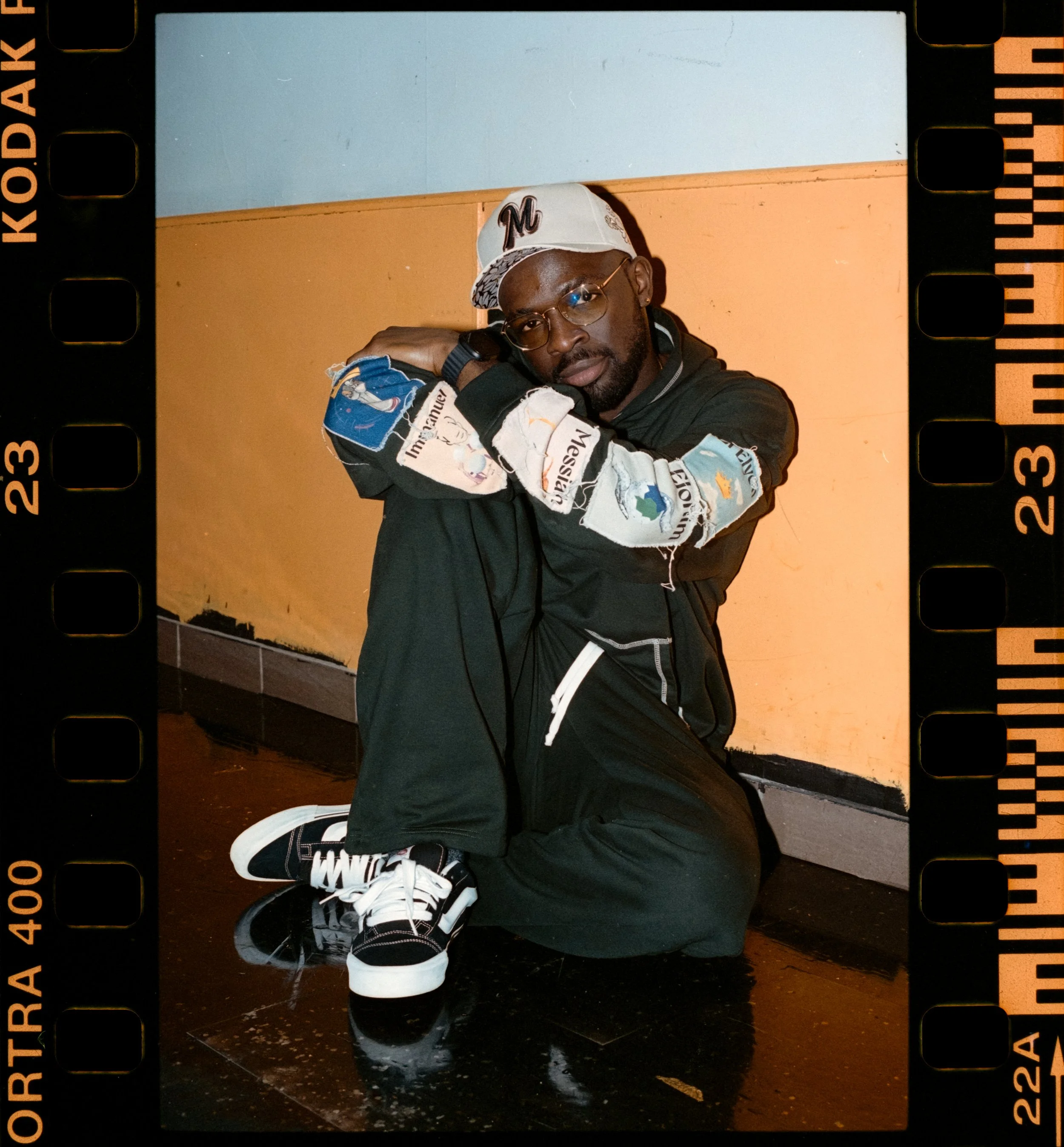 Artist Portrait: "Adeoluwa" A Man Leaning against a yellow wall with his arms crossed wearing a green tracksuit.