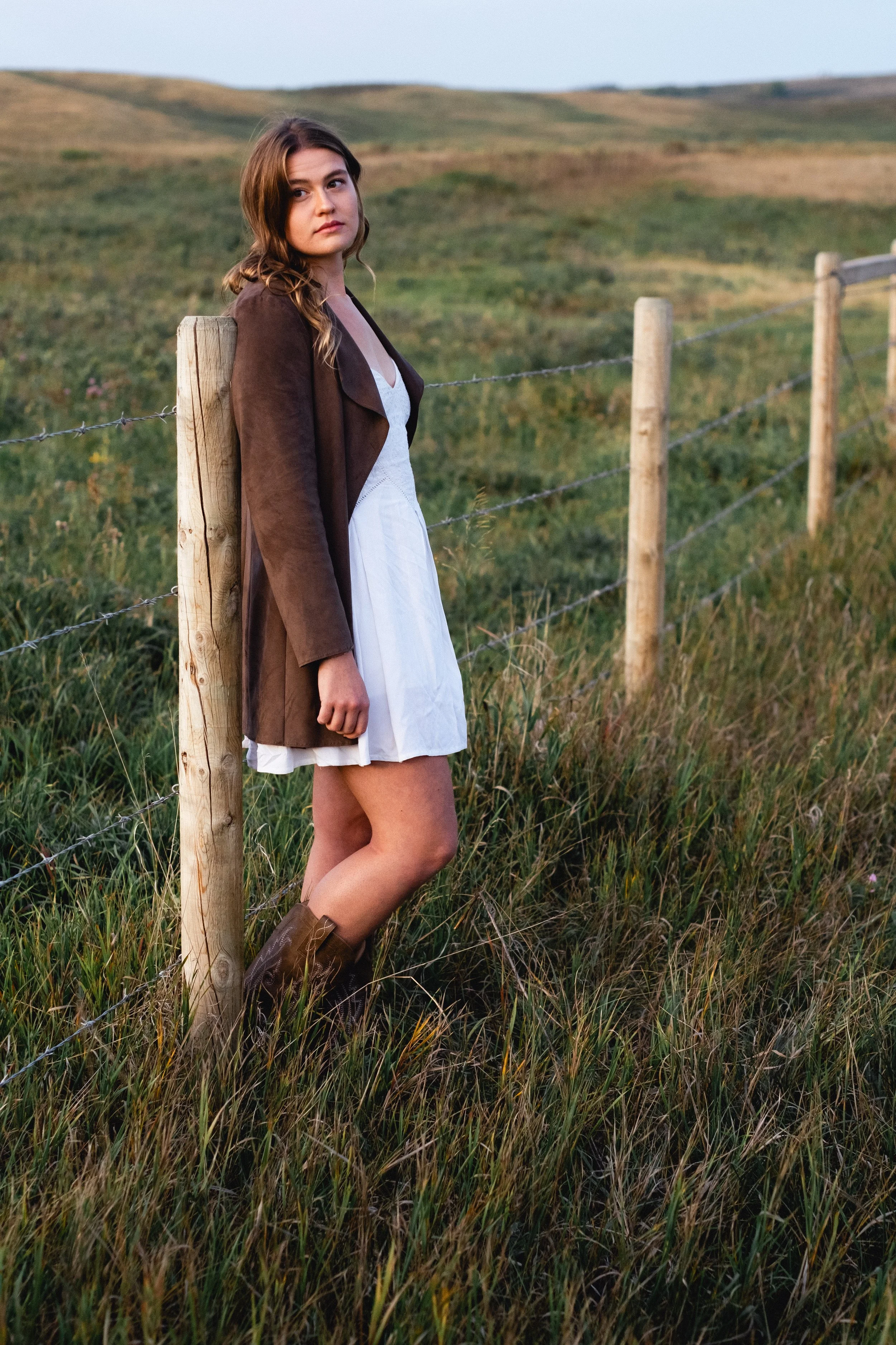 Fashion Photoshoot Calgary. A young woman standing beside a wooden fence in a grassy field during daytime, wearing a brown jacket, white dress, and brown boots.