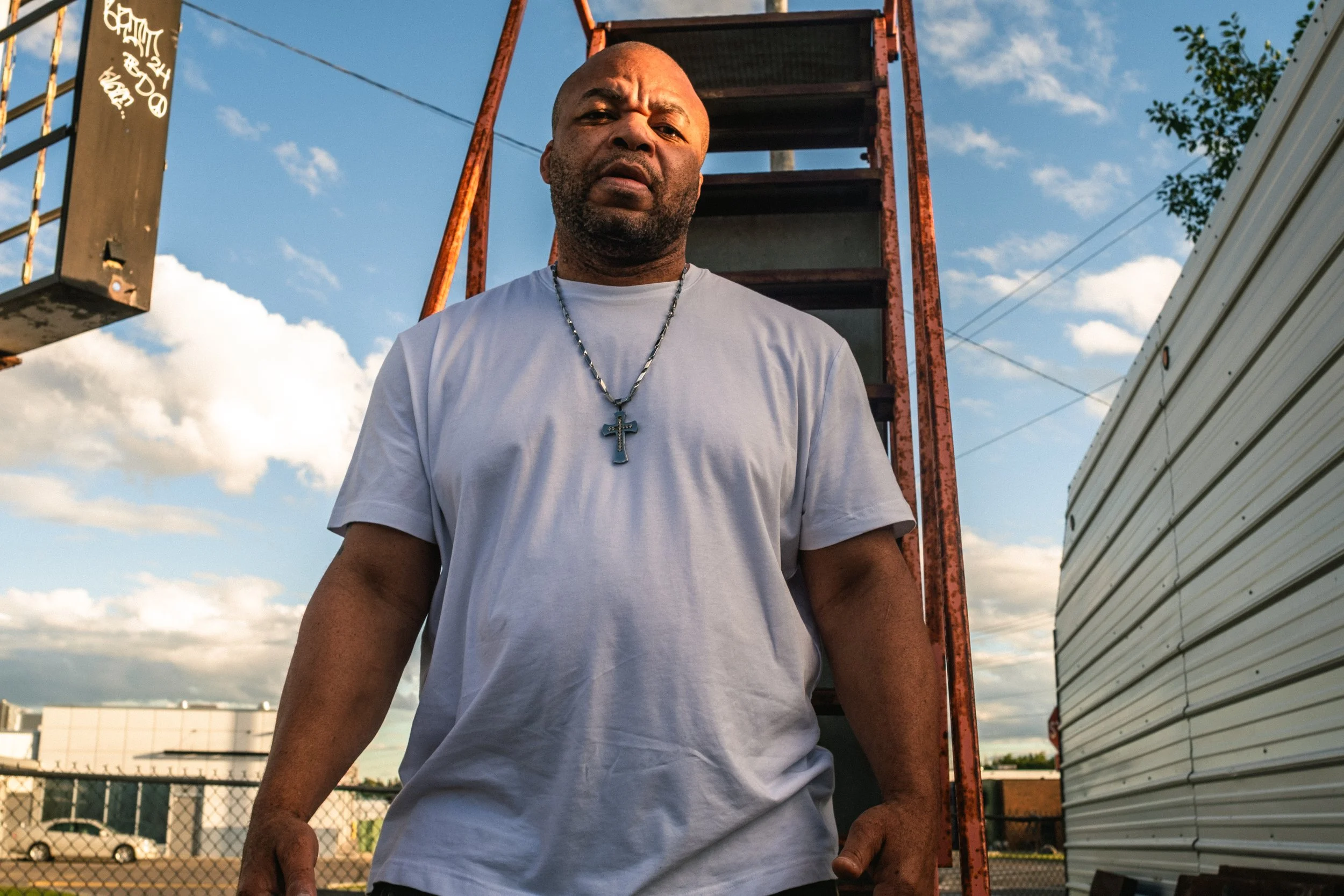 Artist portrait: "Solomon Da Great". A man with a serious expression standing outdoors in front of a rusty fire escape ladder, wearing a white t-shirt and a cross necklace, with a blue sky and clouds in the background.
