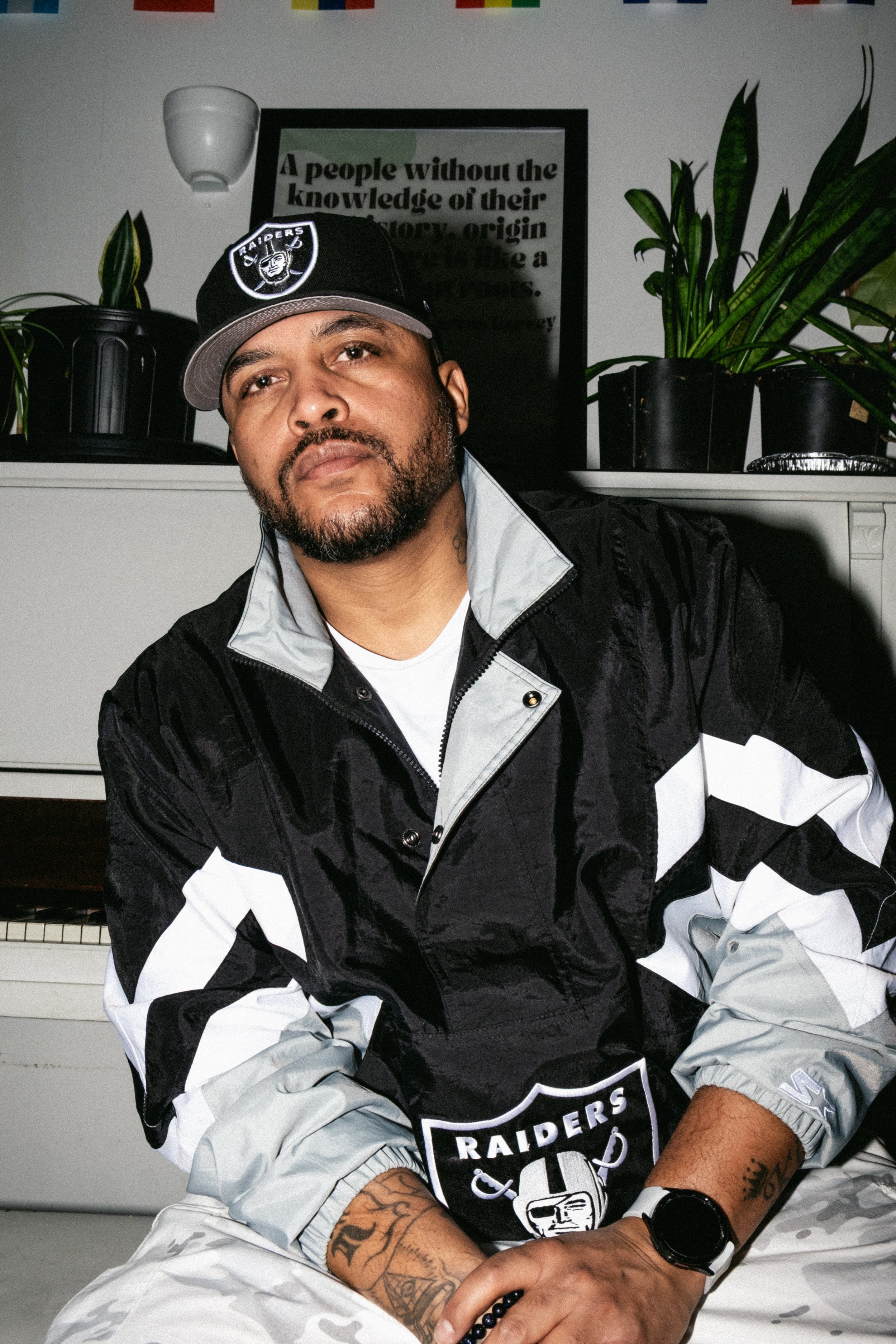 Artist Portrait: "Will Haze" A Man sitting in front of a white piano on the bench wearing a raiders jacket and hat with white camo. Plants and a framed quote on top of the vertical piano in the background. 