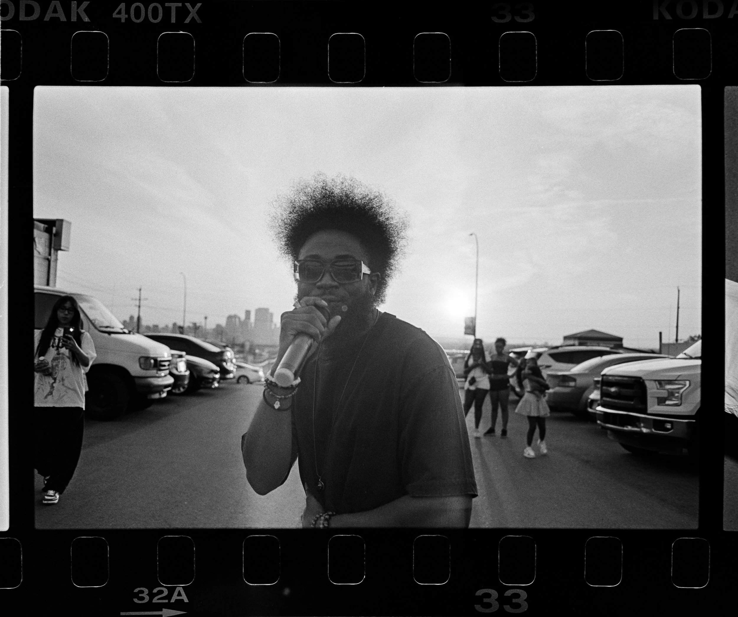 A person with curly hair, sunglasses, and a microphone standing outdoors at a parking lot with cars and a city skyline in the background during sunset.