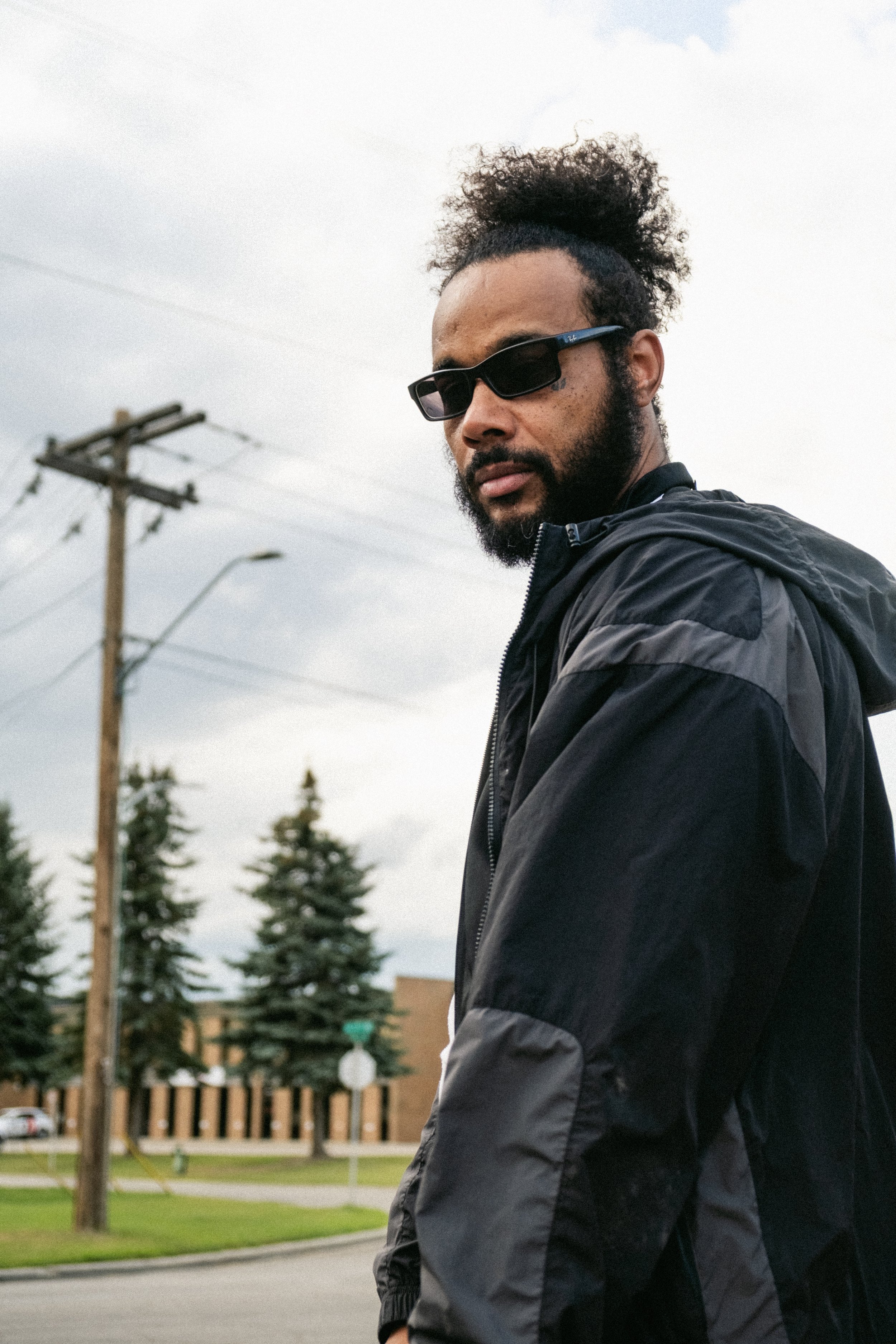 Artist Portrait: "Khanfidence". With curly hair and a beard wearing sunglasses and a black jacket standing outdoors on a cloudy day.