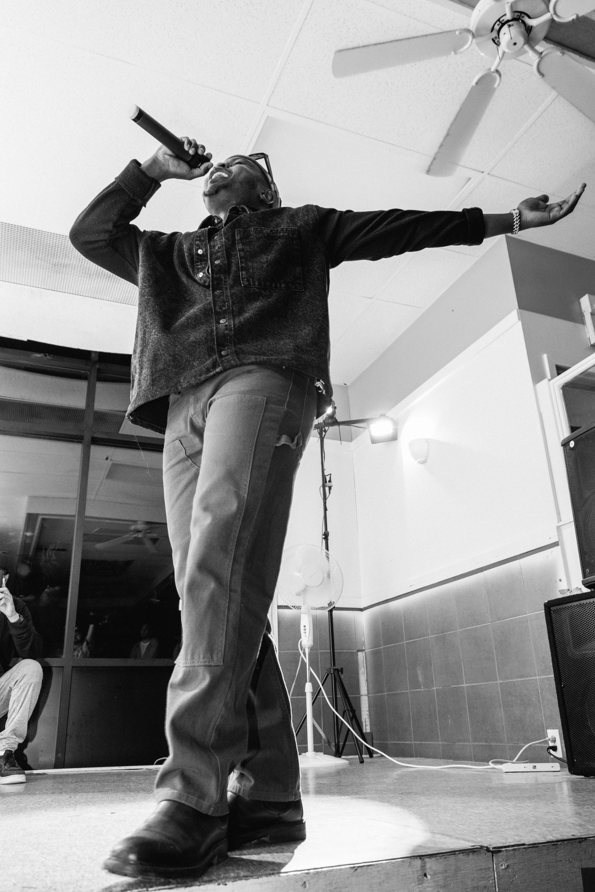 Types singing into a microphone on stage, with one arm extended, in a room with a ceiling fan and speakers.