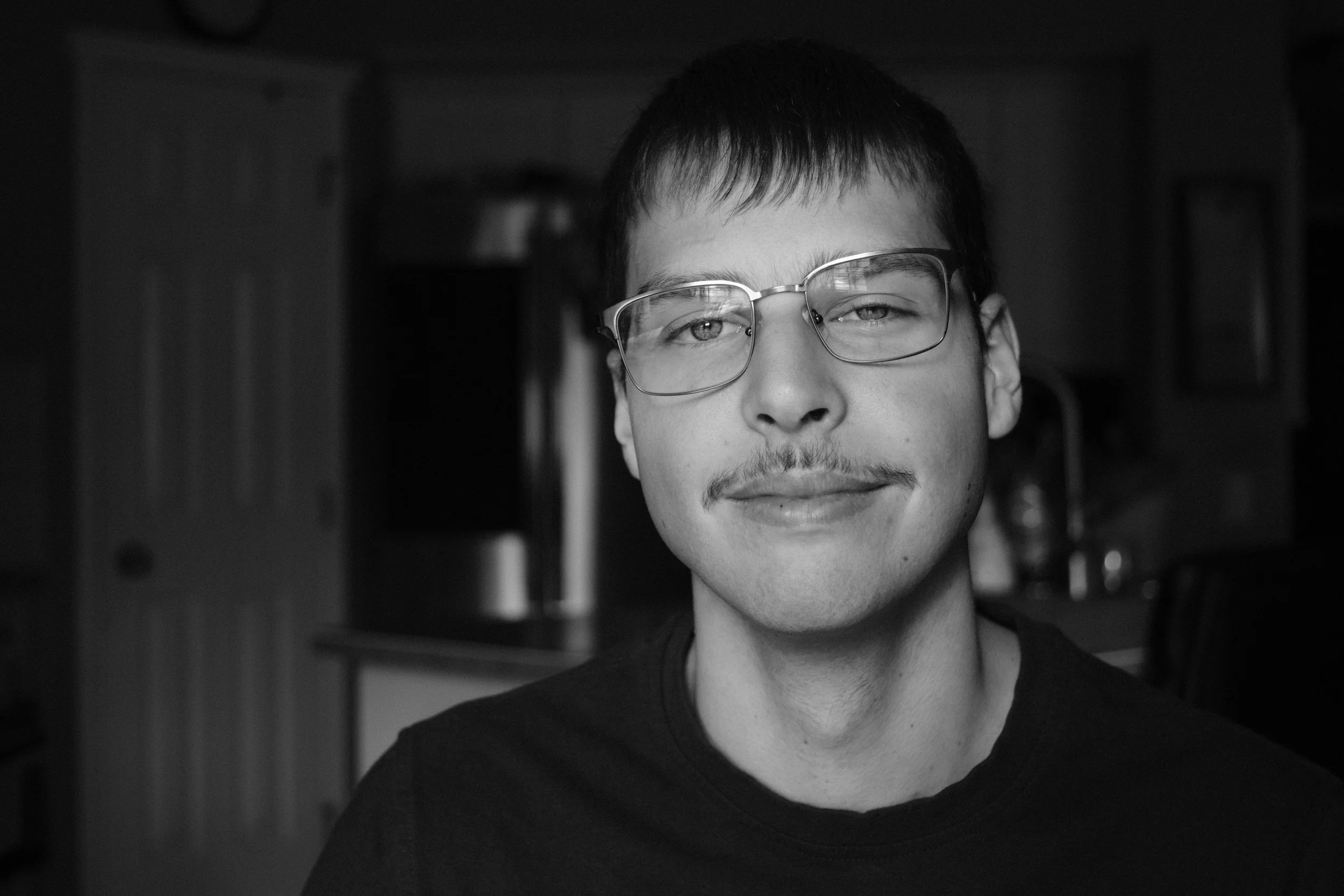 Zachery Knudson, Photographer Portrait. Black and white close-up photo of Zachery Knudson, Photographer with glasses, a faint mustache, and short hair, smiling softly in a room with wooden cabinets in the background.
