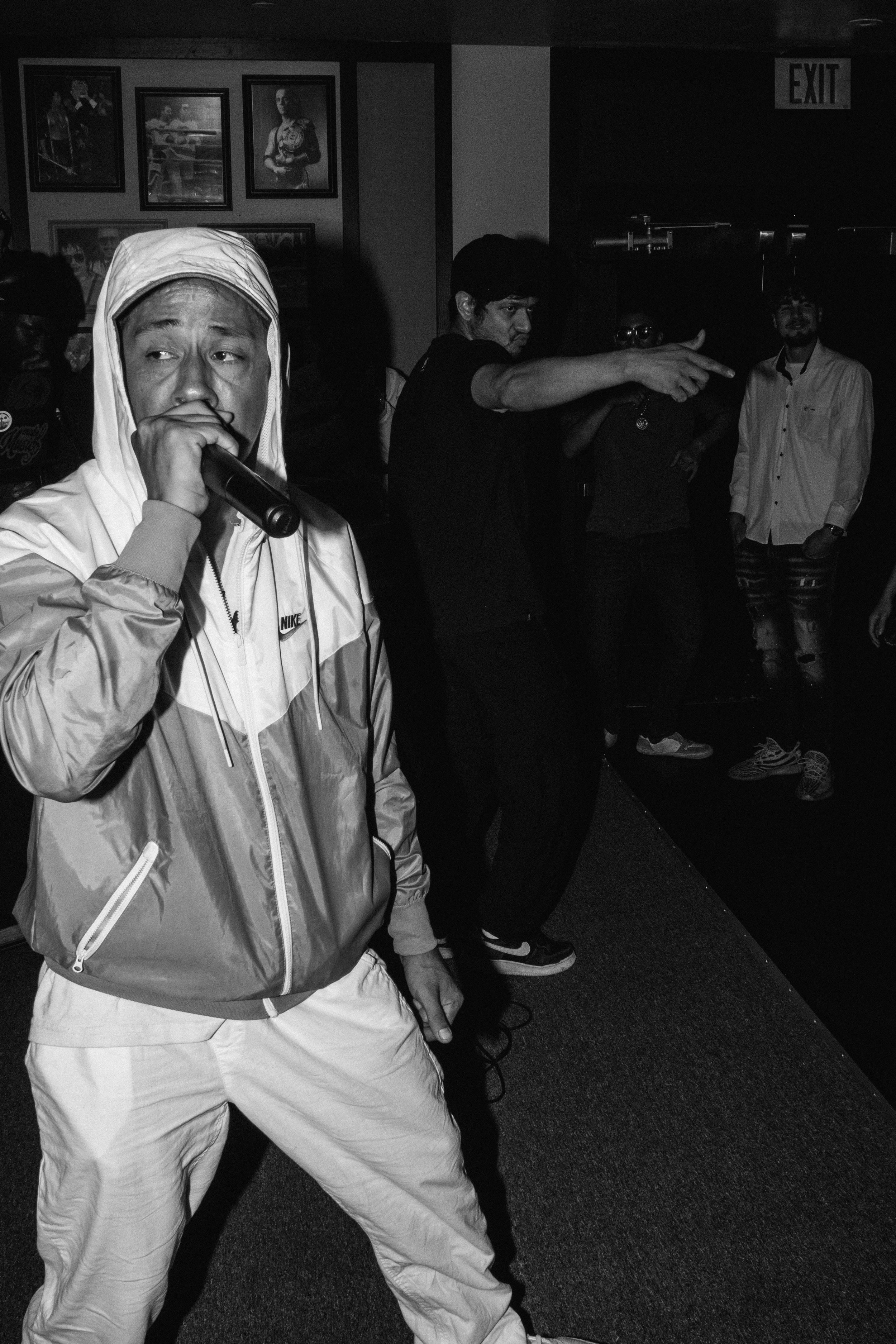 Tecun B'alam Music Performance Photo. in a Nike windbreaker jacket and light-colored pants holding a microphone and singing or speaking into it at a dimly lit indoor event.
